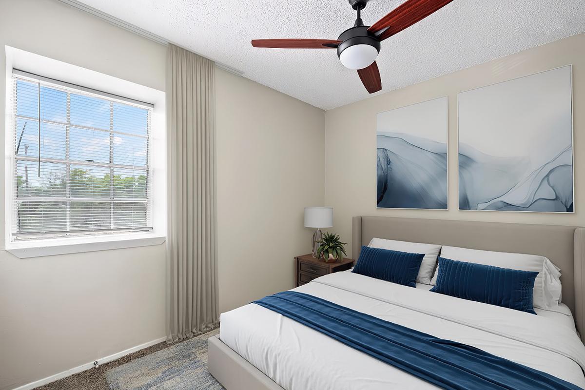 A cozy bedroom featuring a bed with white linens and a blue throw blanket. There are two abstract art pieces on the wall above the bed, a small nightstand with a plant beside it, and a large window with sheer curtains allowing natural light to fill the room. A ceiling fan is visible.