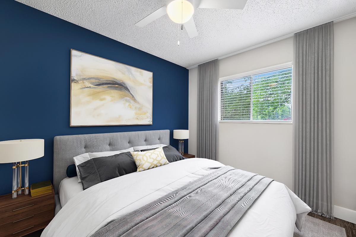 A cozy bedroom featuring a gray upholstered bed with white linens and decorative pillows. A large abstract painting in gold and blue hangs above the bed. Two bedside tables with modern lamps are positioned on either side. A window with sheer curtains allows natural light to enter, illuminating the space.