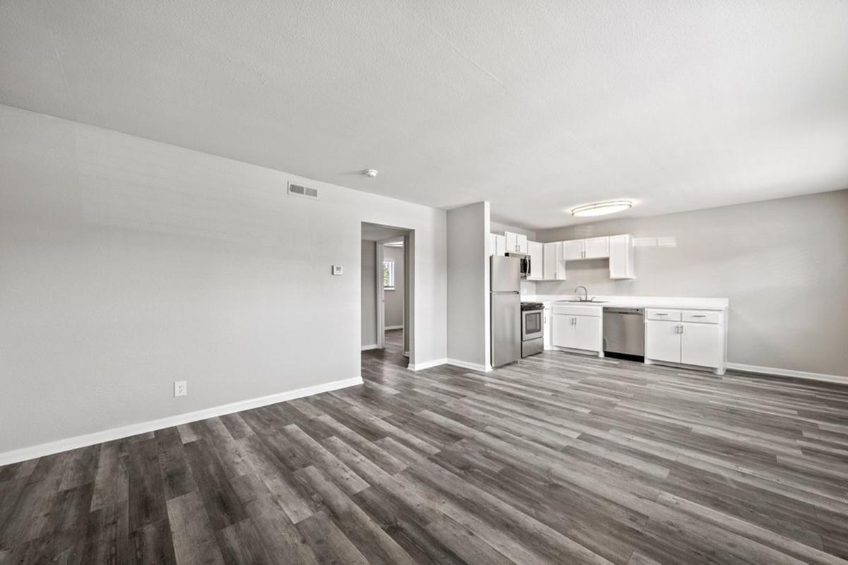 Spacious interior of an apartment featuring an open kitchen with stainless steel appliances, white cabinetry, and a modern light fixture. The living area has large windows and new laminate flooring, creating a bright and inviting atmosphere.