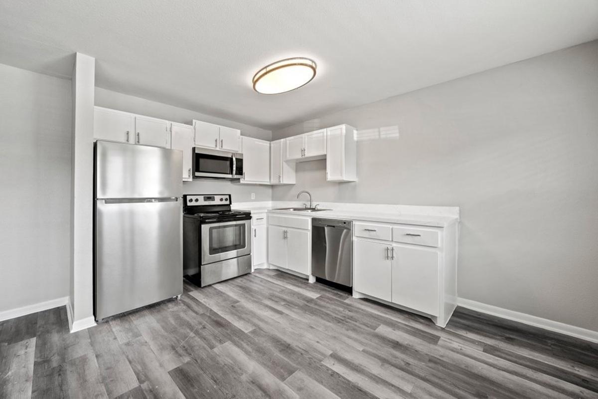 Modern kitchen featuring stainless steel appliances, including a refrigerator, stove, and dishwasher. The cabinets are white, and the countertop is light-colored. The floor is made of wood-like laminate, and there is a circular ceiling light illuminating the space. Walls are painted in a neutral tone, creating a bright, airy atmosphere.