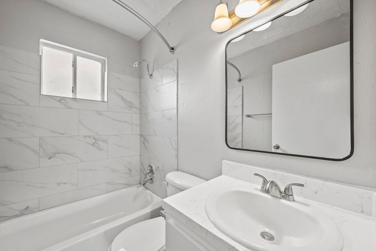 A modern bathroom featuring a bathtub with a shower, a wall-mounted mirror above a white sink, and light-colored marble tiles. A small window allows natural light, and there's a towel rack beside the toilet. The overall design is clean and minimalist.