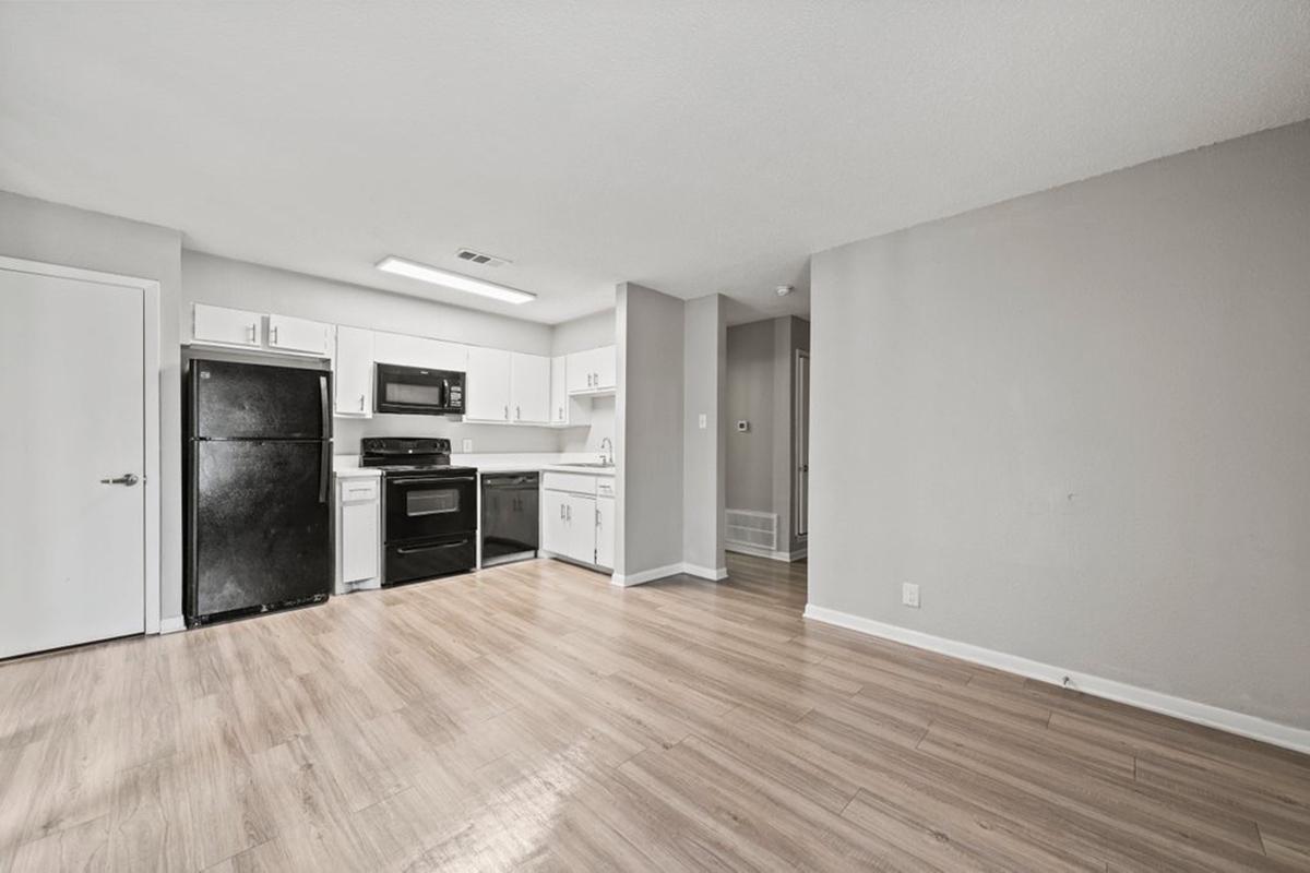 A modern, minimalistic kitchen and living area with light wood flooring. The kitchen features white cabinets, black appliances, and a countertop. On the left, there is a black refrigerator and a matching stove and microwave. The space is well-lit and has neutral-colored walls, creating an open and airy feel.