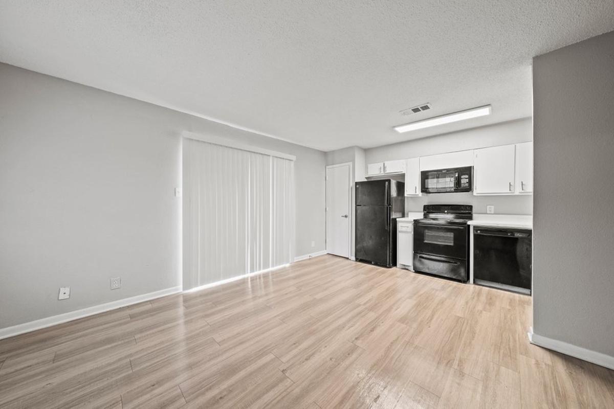 A modern kitchen area featuring light wood flooring, white cabinets, a black refrigerator, a microwave, and an oven. The space has a sliding glass door allowing natural light and minimalistic decor, creating an open and airy atmosphere.