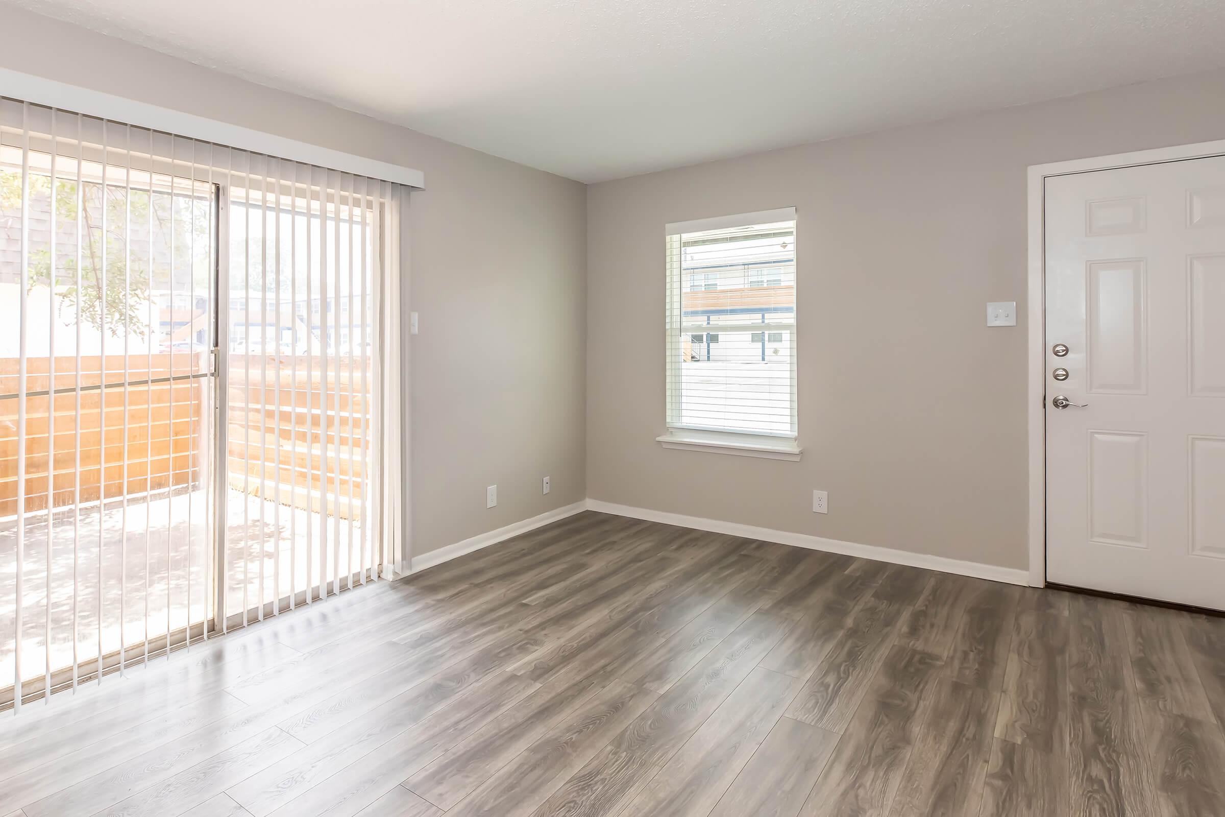 Bright, empty living room with light gray walls and laminate flooring. A sliding glass door with vertical blinds opens to a balcony, while a smaller window allows additional natural light. There's a front door on the right side, adding to the open and inviting space.