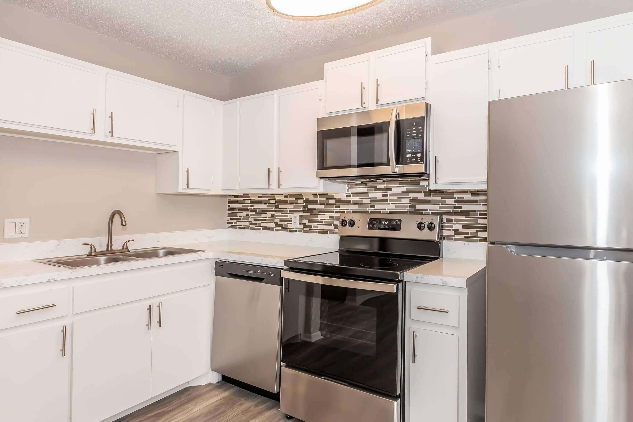 Modern kitchen featuring white cabinetry, stainless steel appliances including a microwave, stove, dishwasher, and refrigerator. The countertop is light-colored, and there is a textured backsplash with small tiles. The kitchen has a clean and contemporary look with ample storage and workspace.
