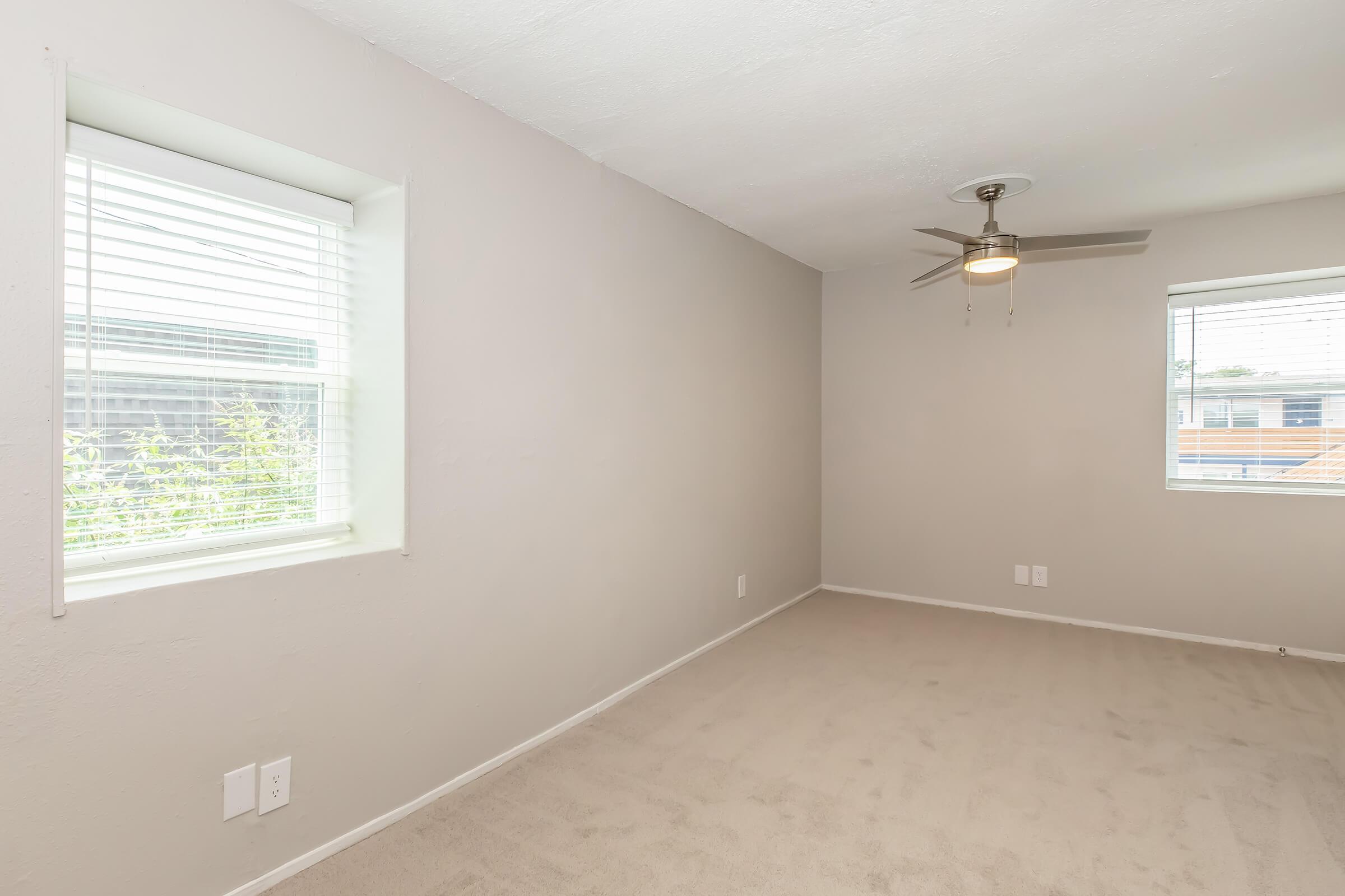 A light, neutral-colored room featuring two windows with blinds, a ceiling fan, and beige carpet flooring. The walls are smooth and unadorned, creating an airy and spacious feel.