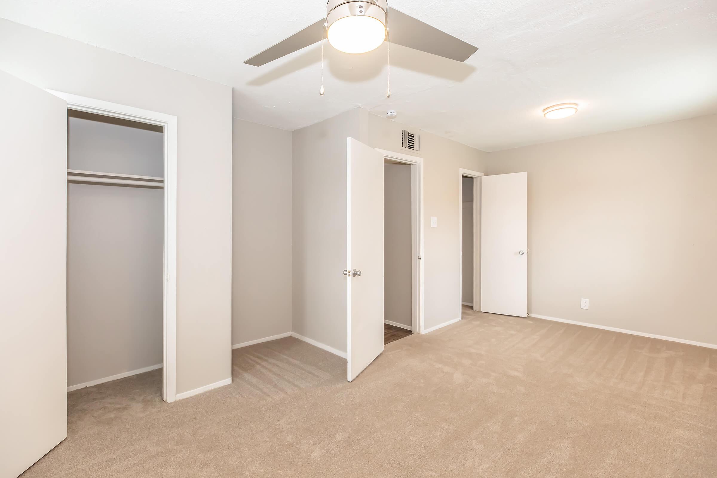 A spacious, neutral-colored bedroom featuring light beige carpet, a ceiling fan, and multiple white doors leading to closets and other areas. The walls are painted gray, adding a modern touch, and the room has ample natural light.