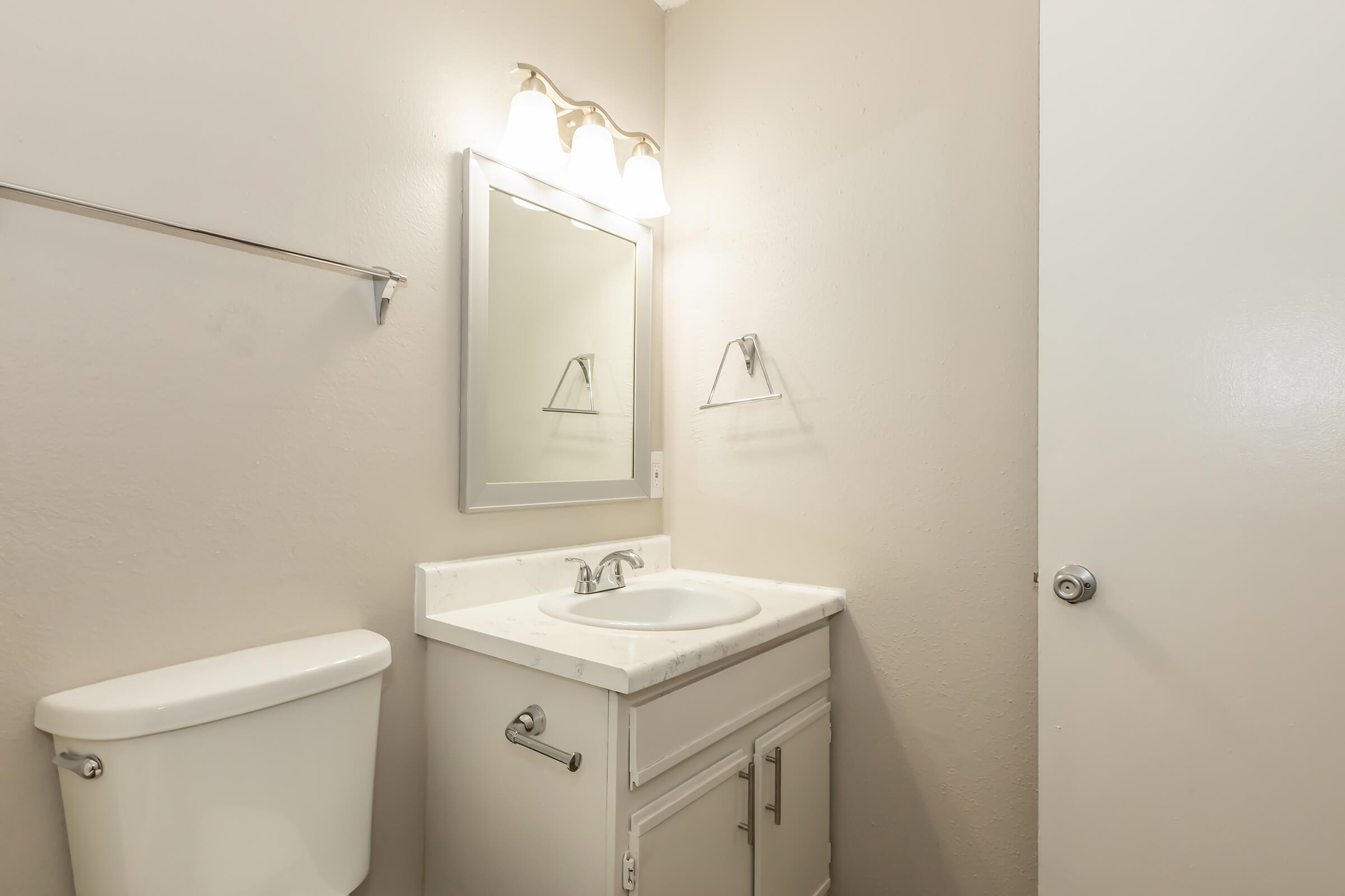 A simple bathroom featuring a white sink with a countertop, a mirror above the sink, a toilet to the left, and a light fixture with three bulbs. The walls are painted in a light color, and there's a towel rack beside the sink. A door is visible on the right side of the image.