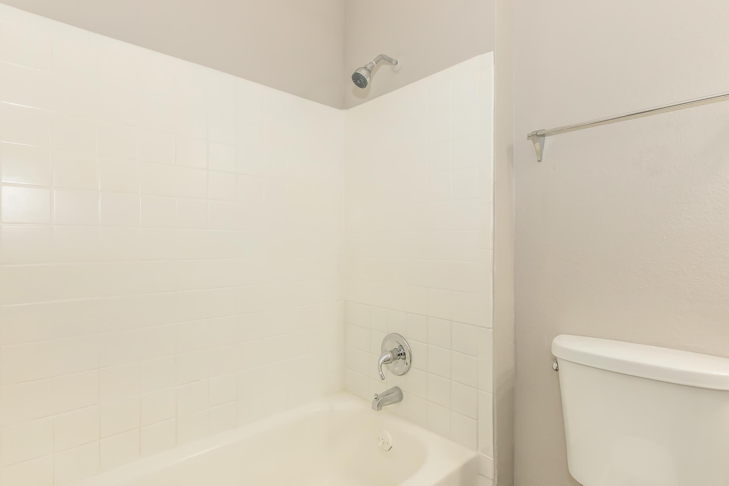 A clean, white tiled bathroom featuring a bathtub with a shower faucet, a wall-mounted showerhead, and a toilet nearby. The walls are painted in a light color, creating an open and bright atmosphere.