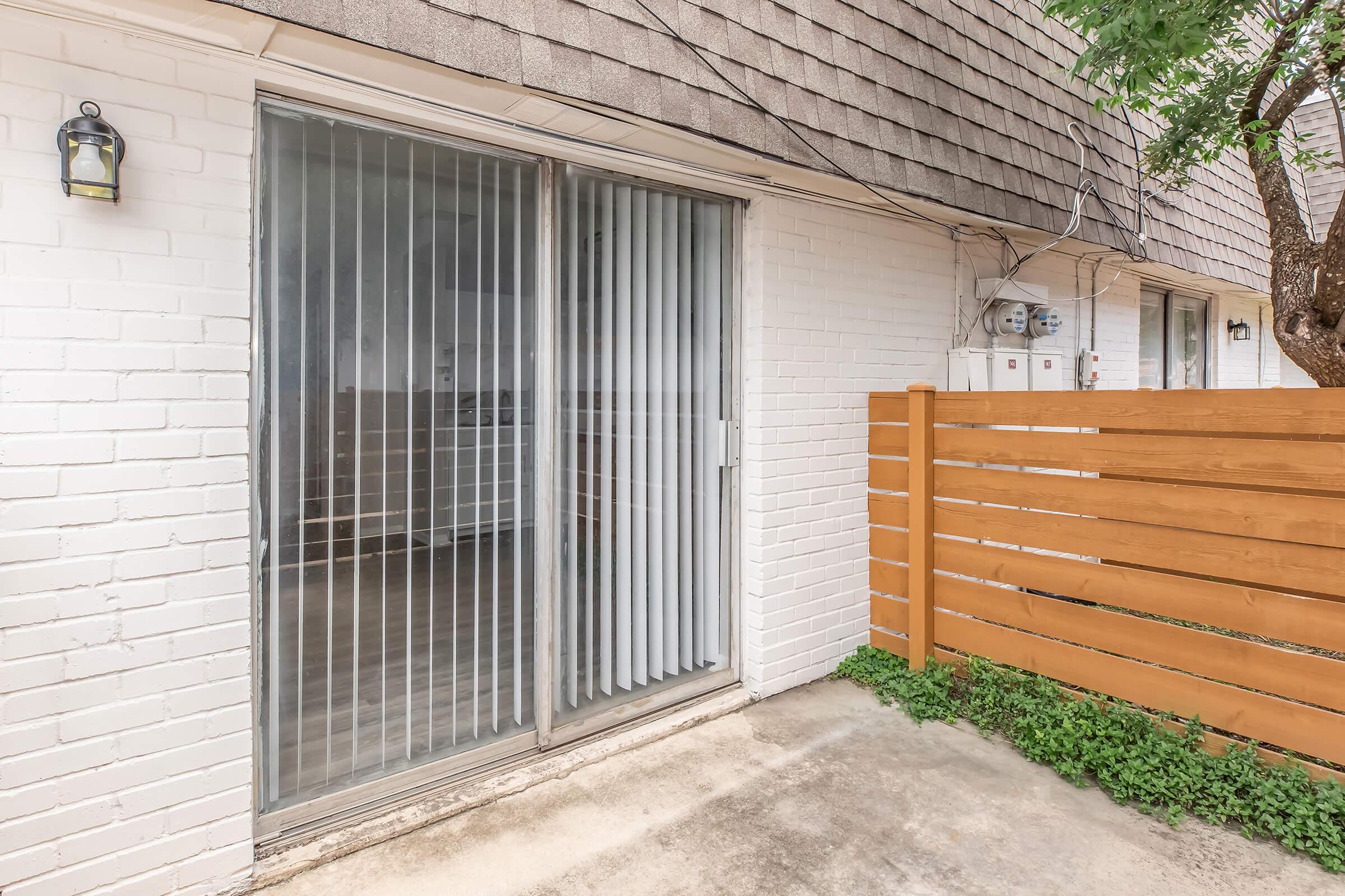 Exterior view of a sliding glass door with vertical blinds, flanked by white brick walls. A wooden fence runs along the side, with greenery at the base. The area appears well-lit and clean, suggesting a residential setting.