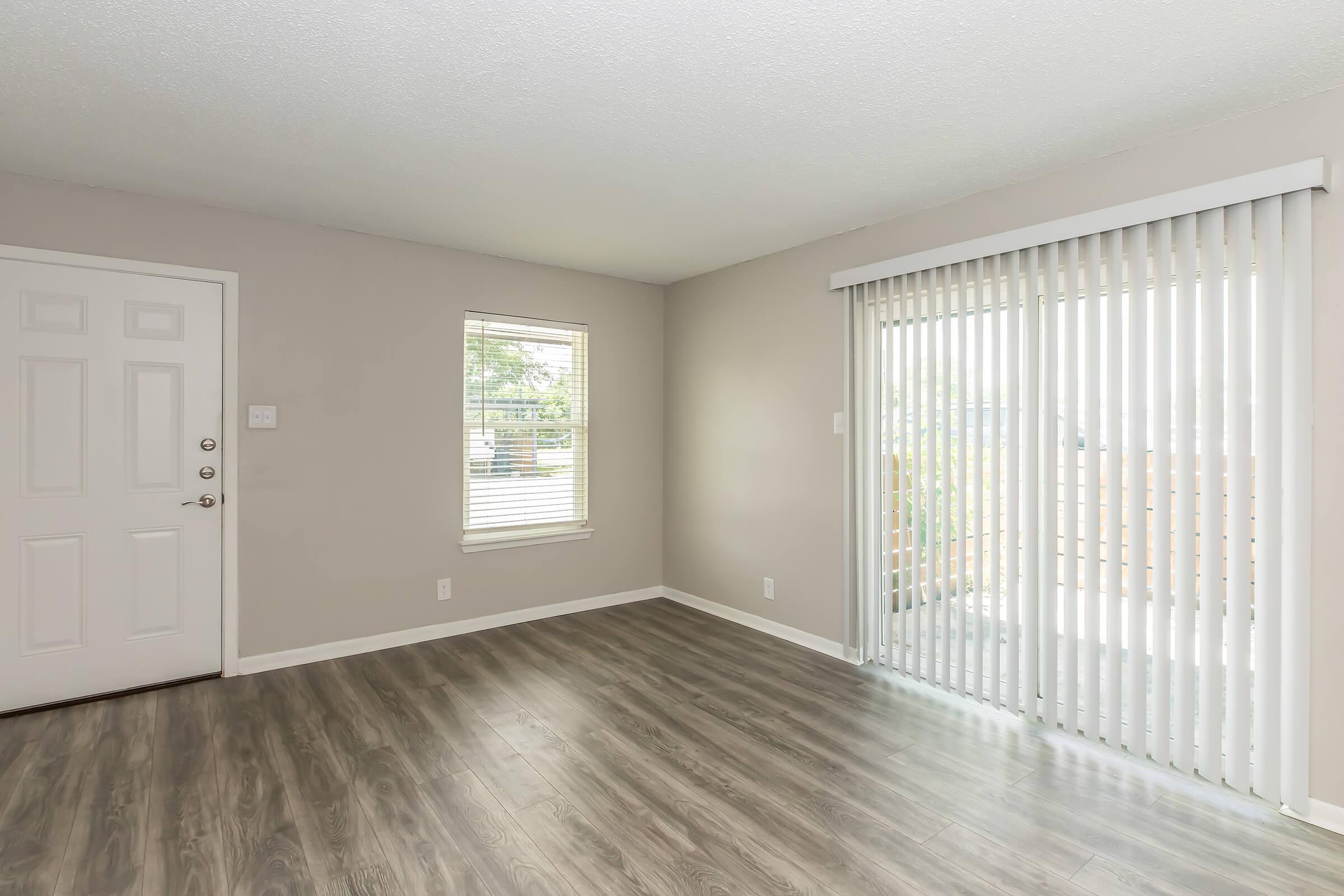 A light and airy room with a gray wall, featuring a front door on the left, a small window with horizontal blinds, and a sliding glass door with vertical blinds leading to an outdoor space. The floor is finished with light wood-like laminate.