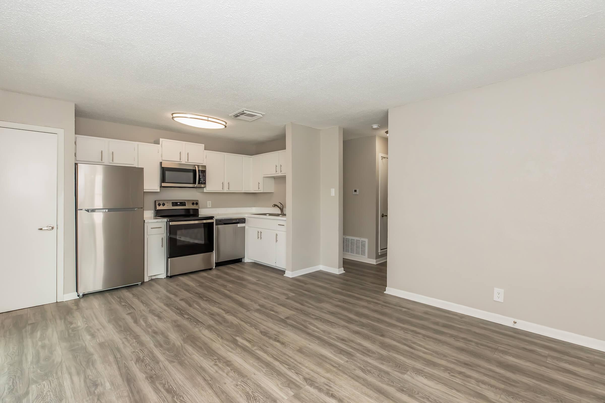 A modern kitchen-living area featuring stainless steel appliances, including a refrigerator and oven, with white cabinetry. The walls are a light neutral color, and the flooring is a wood-like laminate. Bright lighting comes from a circular ceiling fixture, creating a spacious and inviting atmosphere.