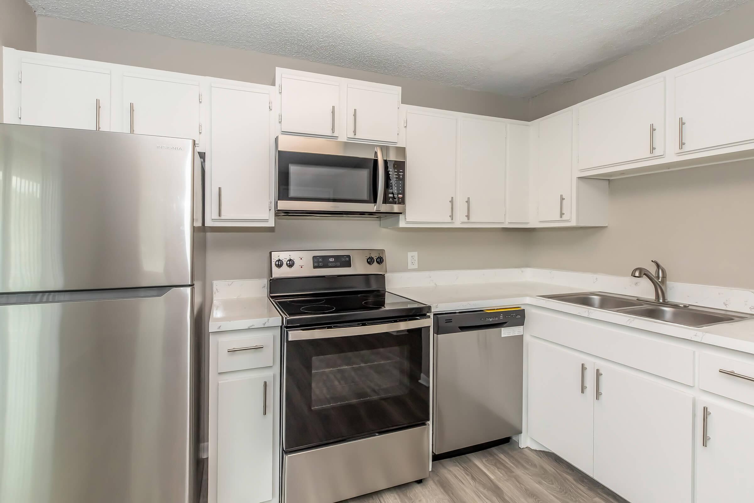 Modern kitchen with white cabinets, stainless steel appliances including a refrigerator, microwave, oven, and dishwasher. The countertop is light-colored, and there are dual sinks with a window above. The flooring is light wood-like laminate.