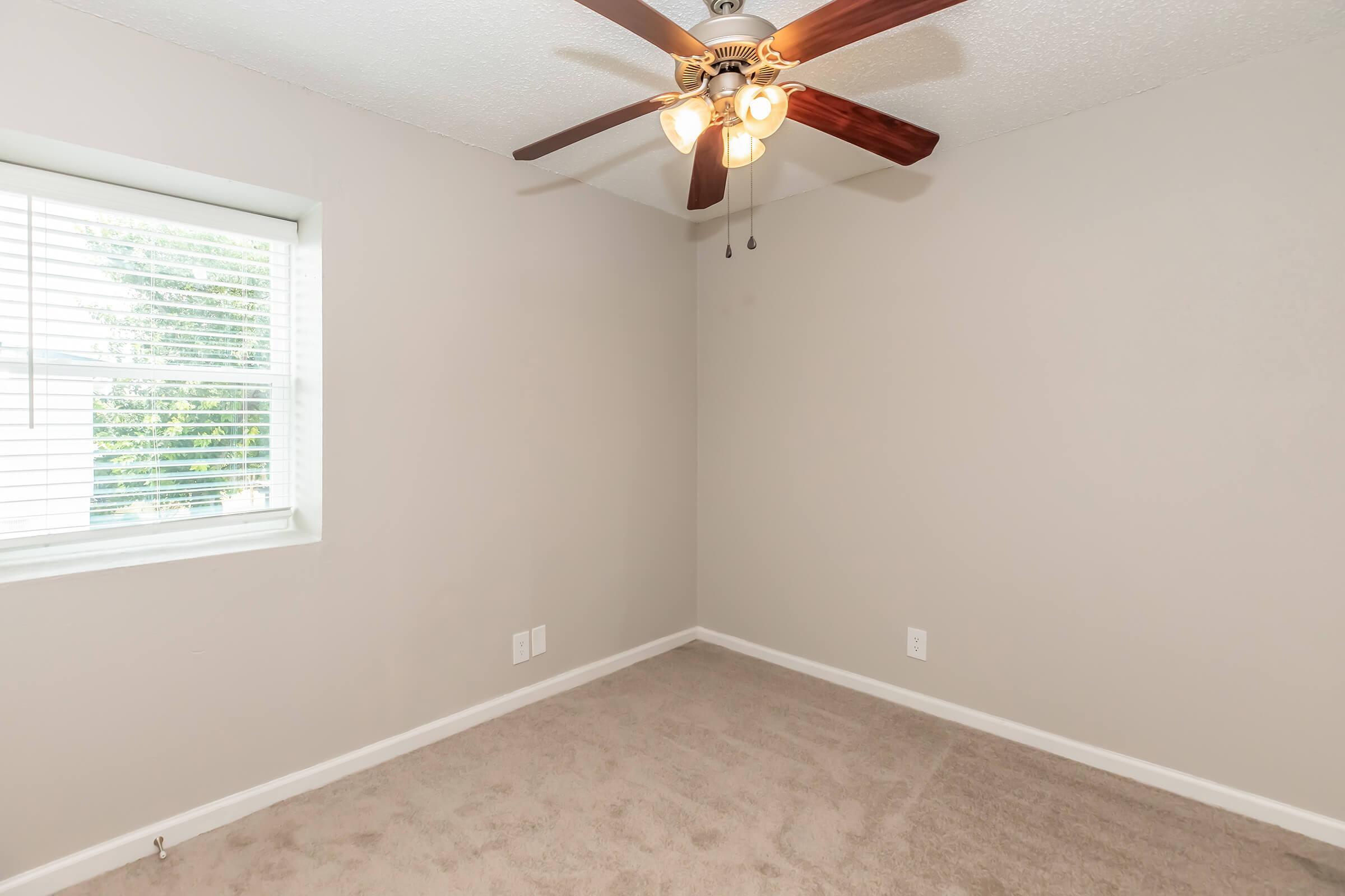 Empty room with light gray walls and beige carpet, featuring a ceiling fan with four bulbs. A window with white blinds allows natural light to enter, illuminating the space. The room has minimal furnishings and clean lines, making it look spacious and well-lit.