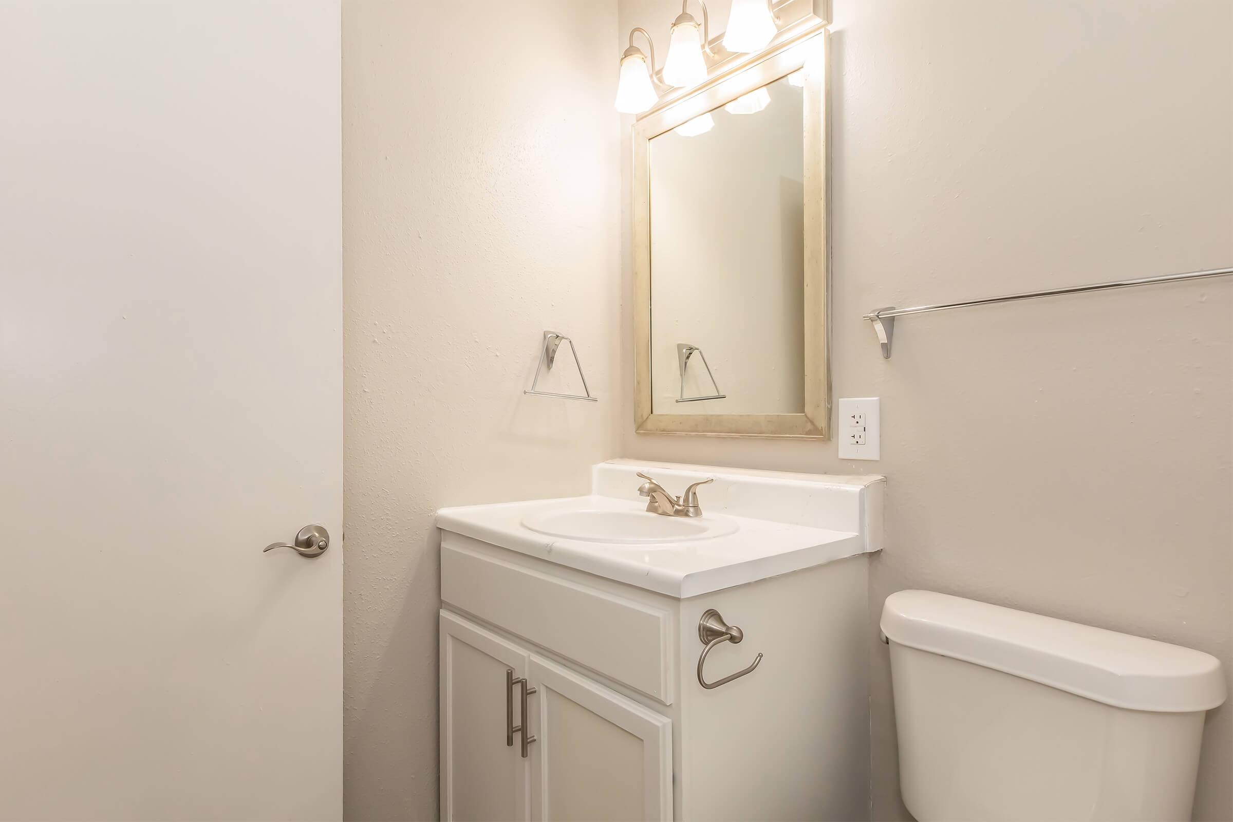 A simple bathroom featuring a white sink with a mirror above, a light fixture with three bulbs, a toilet, and a wall-mounted towel rack. The walls are painted light gray, creating a clean and modern aesthetic. A white door leads to the room.