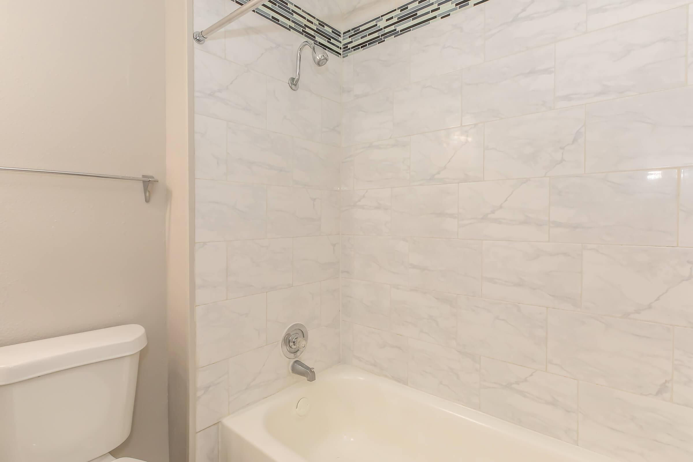 A clean, modern bathroom featuring a white bathtub with a showerhead above, surrounded by light marble tiles. The wall has a decorative accent strip at the top, and a white toilet is visible to the left. The overall aesthetic is bright and minimalist.