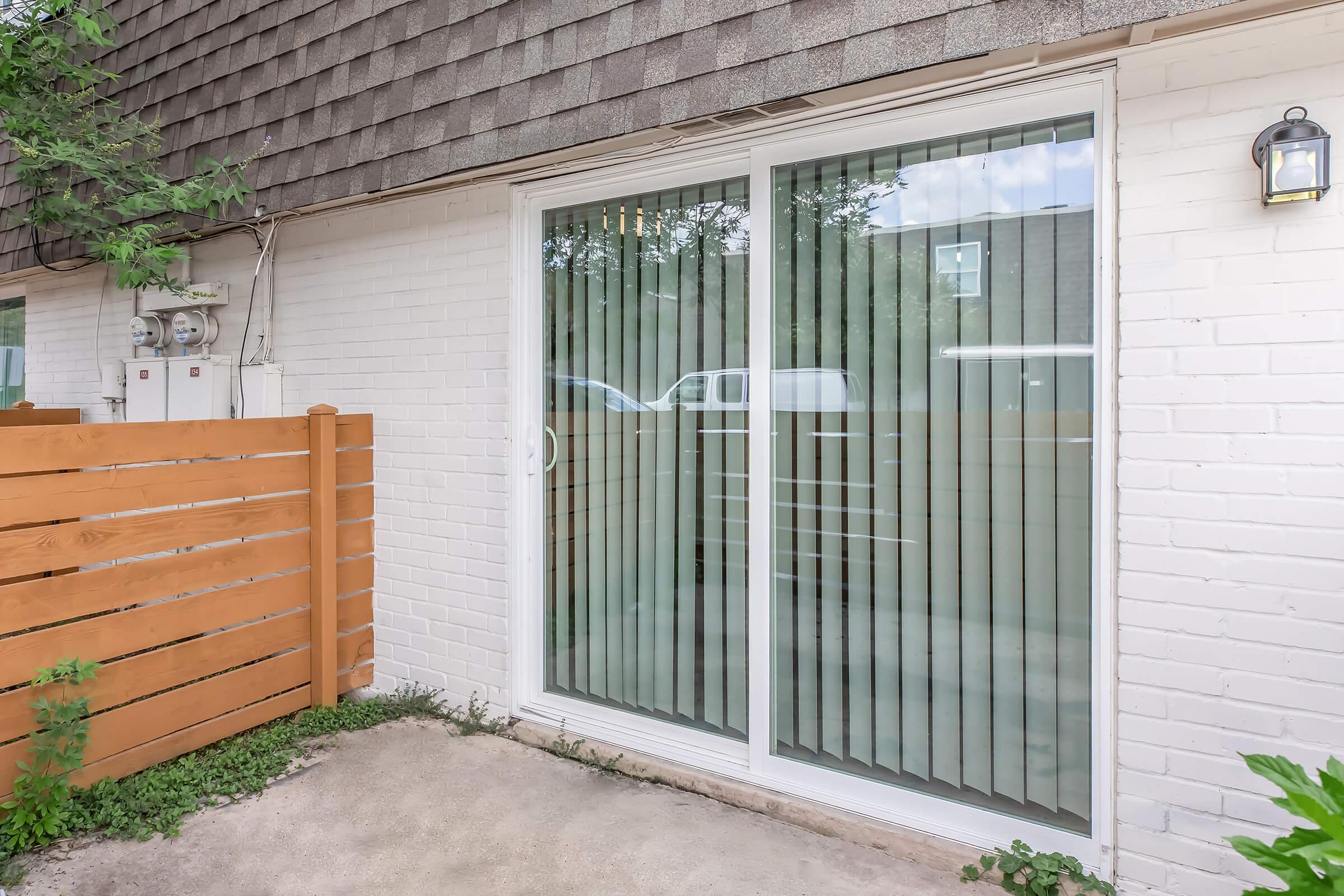 Sliding glass doors with vertical blinds open to a small outdoor area. A wooden fence frames the space, and nearby greenery adds a natural touch. Visible utilities are situated to the left of the doors. The overall setting appears to be a residential patio or backyard.