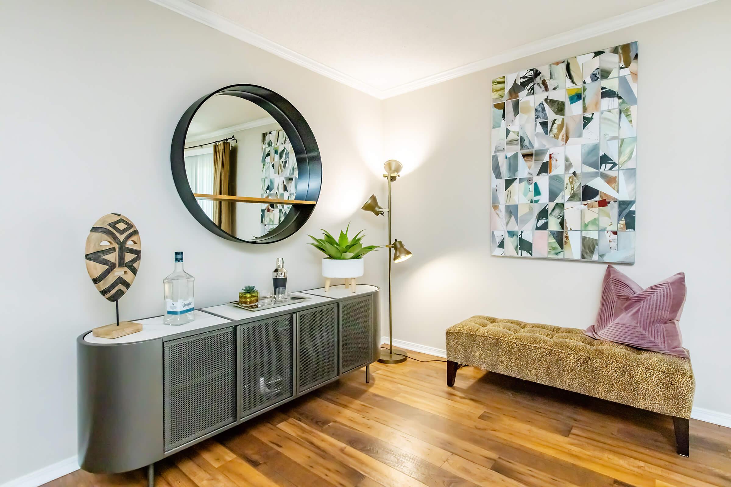 A stylish living room corner featuring a round mirror above a modern console table with decorative items, including a plant and a bottle. There is a cozy animal-print bench and an abstract artwork on the wall, set against light-colored walls and wooden flooring, creating a contemporary and inviting atmosphere.