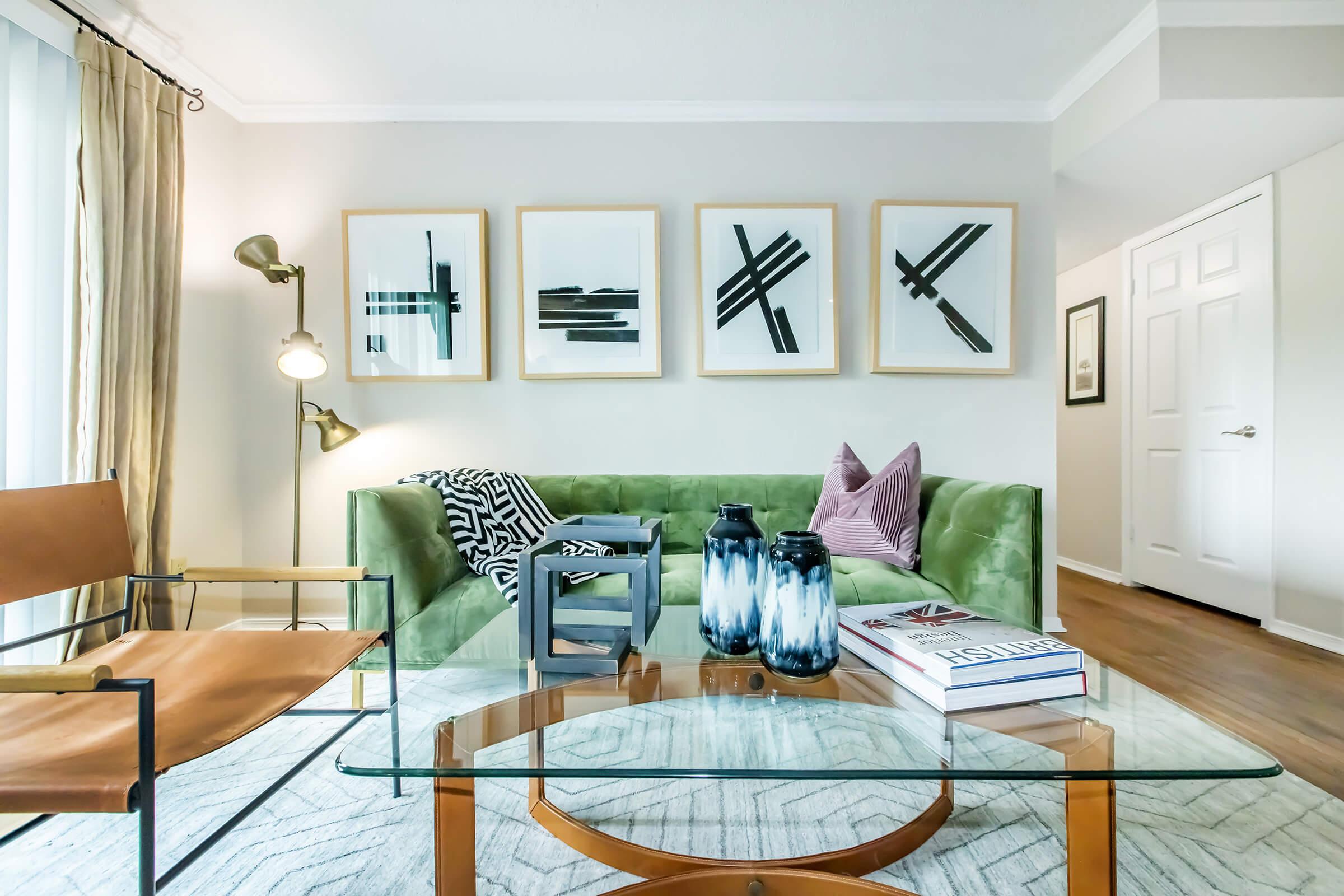A modern living room featuring a green sofa with patterned pillows, a glass coffee table, and a light-colored rug. On the wall, there are five framed abstract artworks. A stylish armchair and a floor lamp complement the space, creating a cozy and contemporary atmosphere.