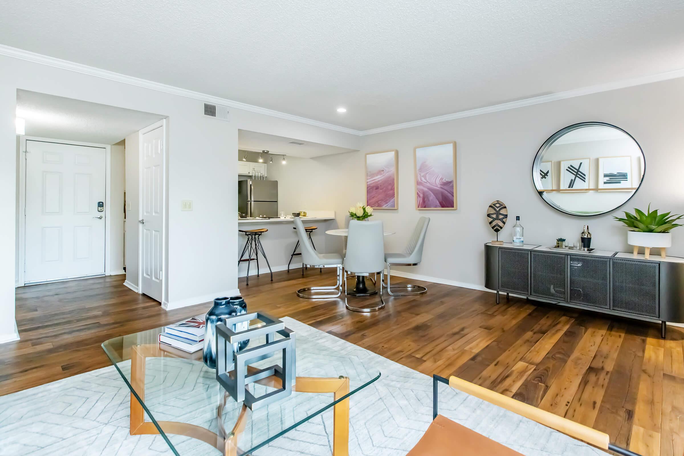 A well-lit modern living room featuring a glass coffee table, comfortable seating, and a stylish sideboard. Artwork adorns the walls, and there are plants for a touch of greenery. The space opens to a kitchen area, with a door leading to the hallway, creating a welcoming atmosphere.