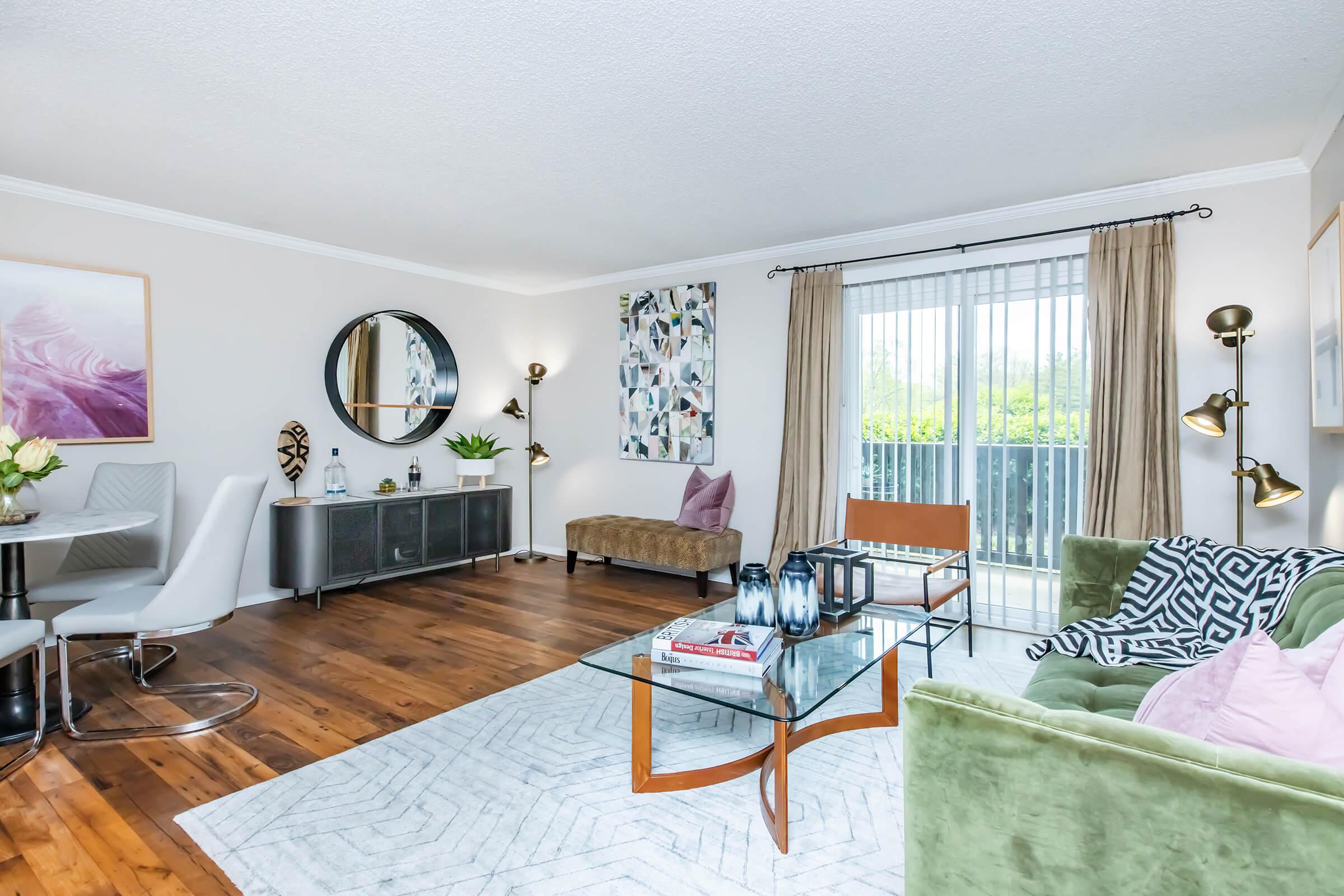 A modern living room featuring a green couch, a glass coffee table, and a chic armchair. Elegant decor includes a round mirror, framed artwork, and stylish lamps. Natural light streams in from sliding glass doors, with wood flooring and a geometric rug creating a welcoming ambiance.