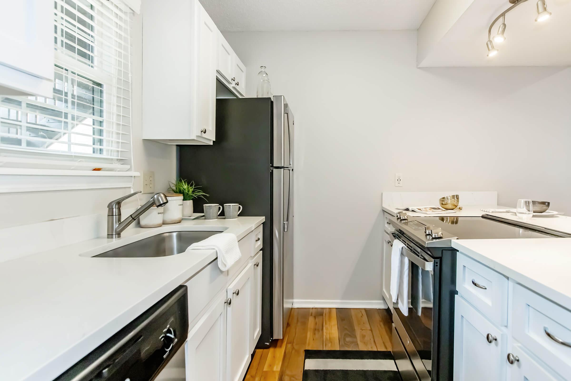 Modern kitchen with white cabinetry, stainless steel appliances, and a clean countertop. Features a sink, a stove, and a refrigerator, along with a few decorative items like mugs and a plant. The flooring is wood, and there is natural light coming through a window.