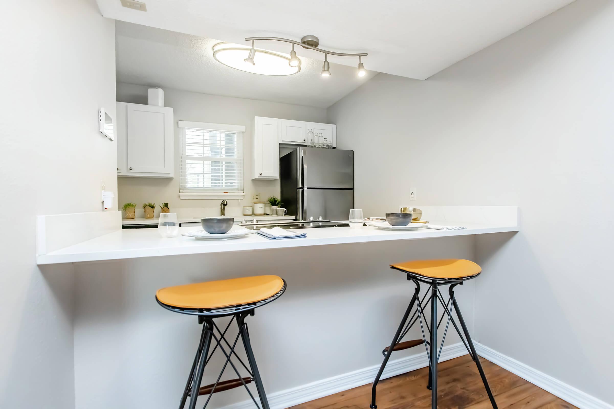 A modern kitchen with a white countertop and two orange bar stools. Stainless steel appliances, including a refrigerator, are visible in the background. The kitchen features white cabinets and a small window with natural light. Minimalistic decor adds a clean and contemporary feel.