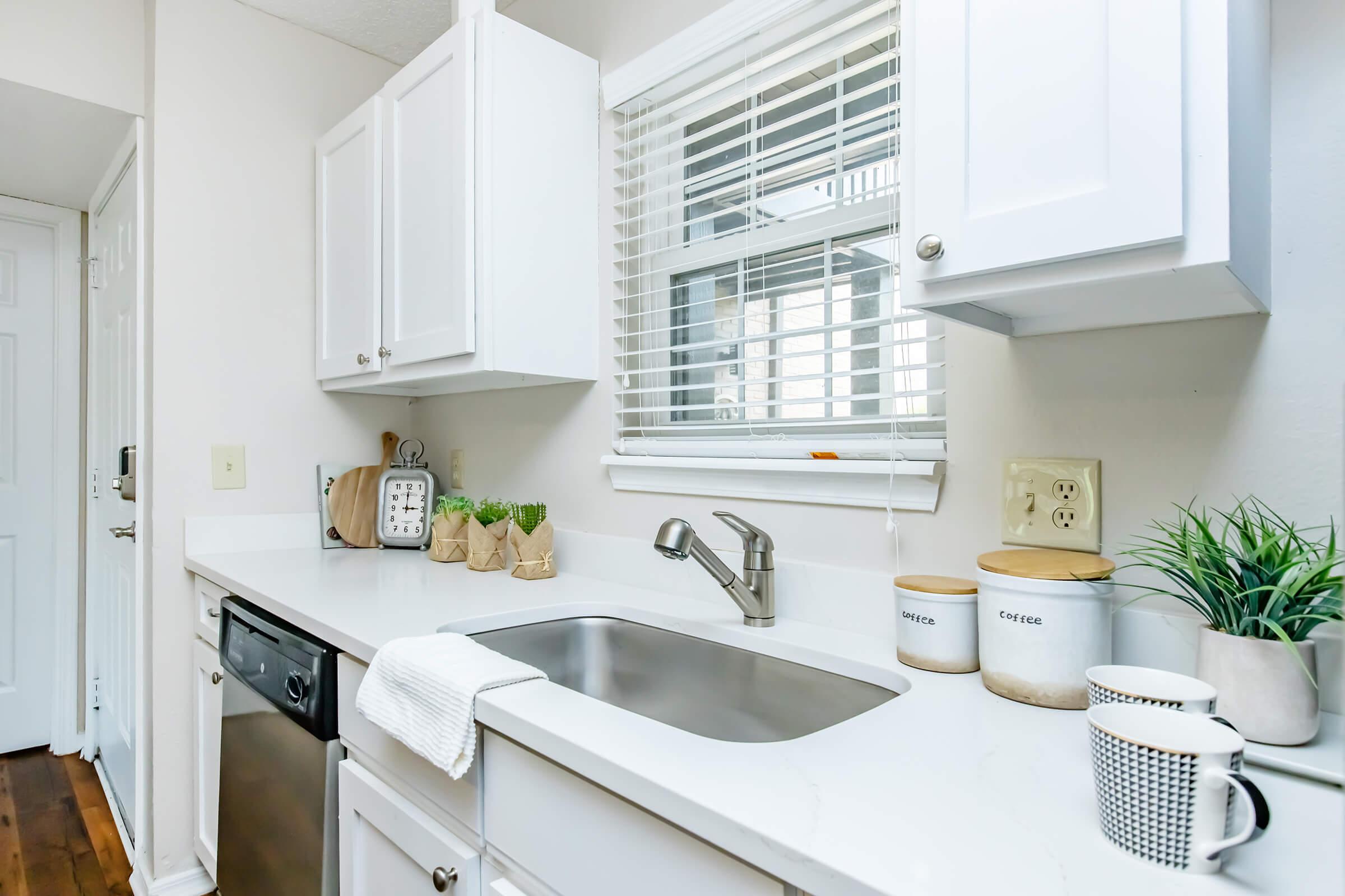 A modern kitchen featuring white cabinets, a stainless steel sink, and a countertop with a small potted plant and canisters. Sunlight streams through a window with blinds, illuminating the clean and tidy space. A dish towel hangs from the sink, and a dishwasher is visible nearby.