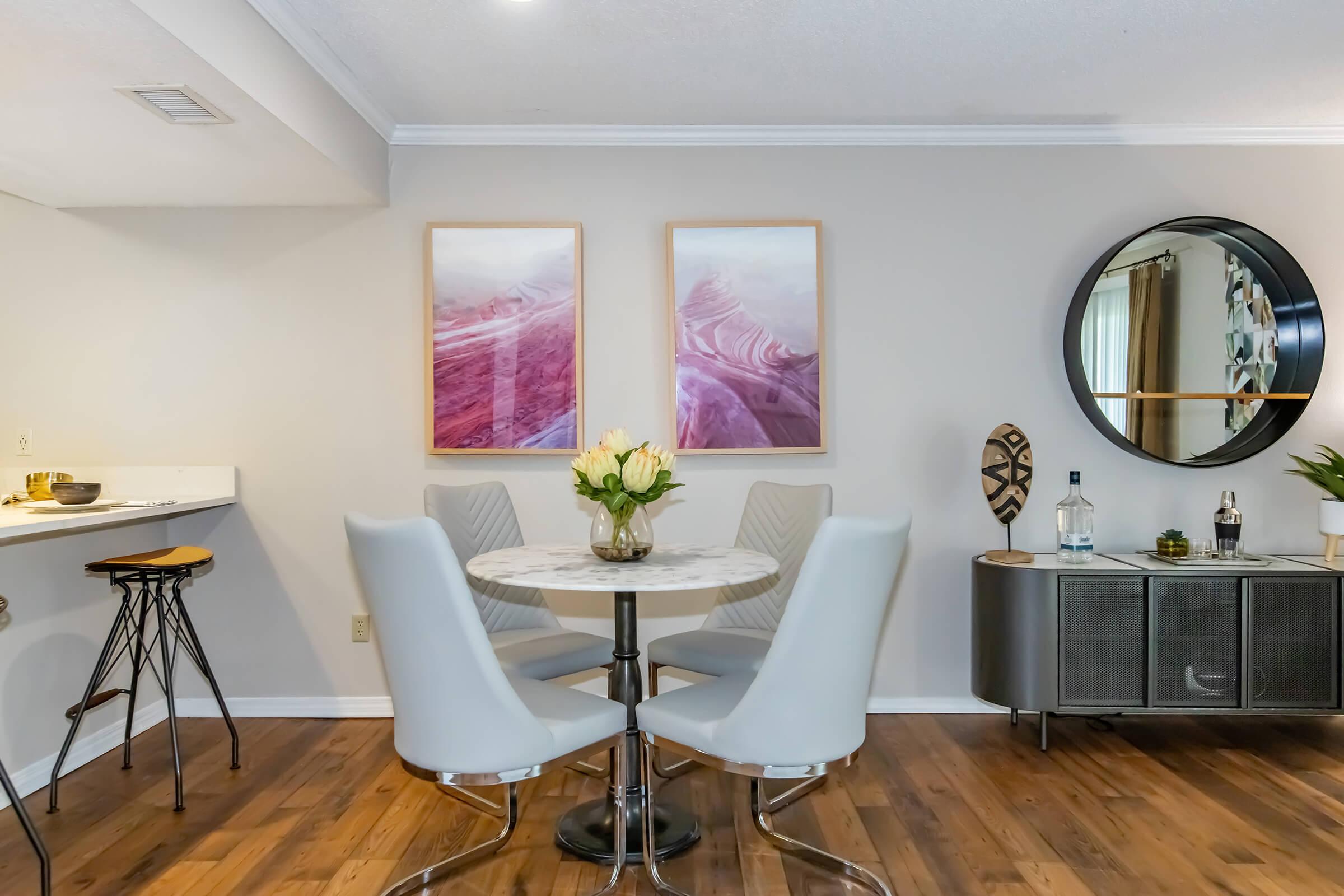 A modern dining area featuring a round table with four white chairs. The wall has two framed abstract artworks in soft pink hues. A circular mirror is mounted nearby, reflecting light. A sideboard holds a decorative bottle and plant, adding a touch of elegance to the space.
