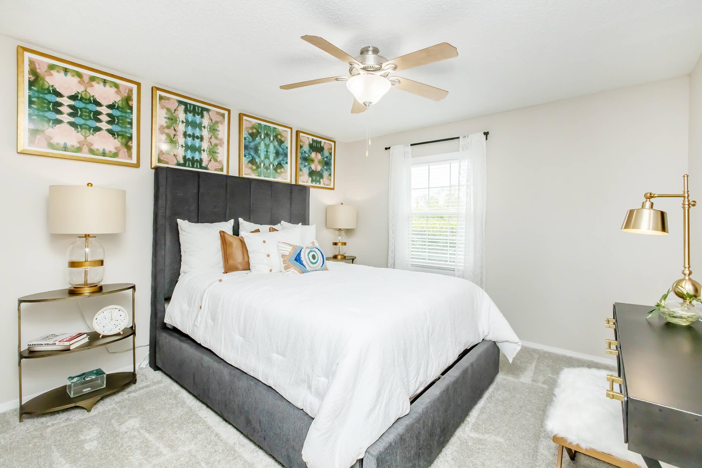 A stylish bedroom featuring a gray upholstered bed with white bedding, decorative pillows, and framed artwork above the headboard. There's a nightstand with a lamp and a clock on one side, and a gold accent table with a lamp on the other. A ceiling fan and bright natural light from a window add to the inviting atmosphere.