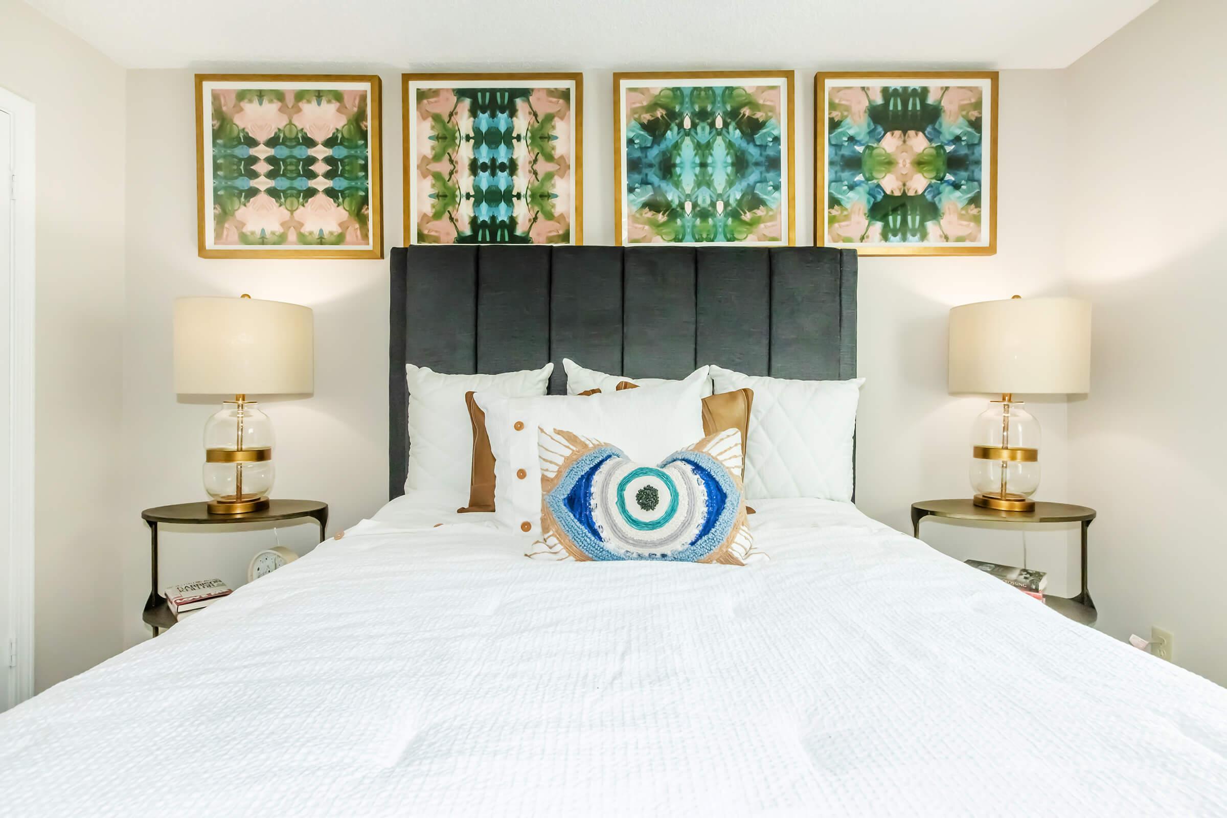 A modern bedroom featuring a plush bed with white bedding and decorative pillows. Above the headboard, there are four framed abstract artworks. On either side of the bed, there are bedside tables with stylish lamps that have a gold base and clear shades, creating a cozy atmosphere.