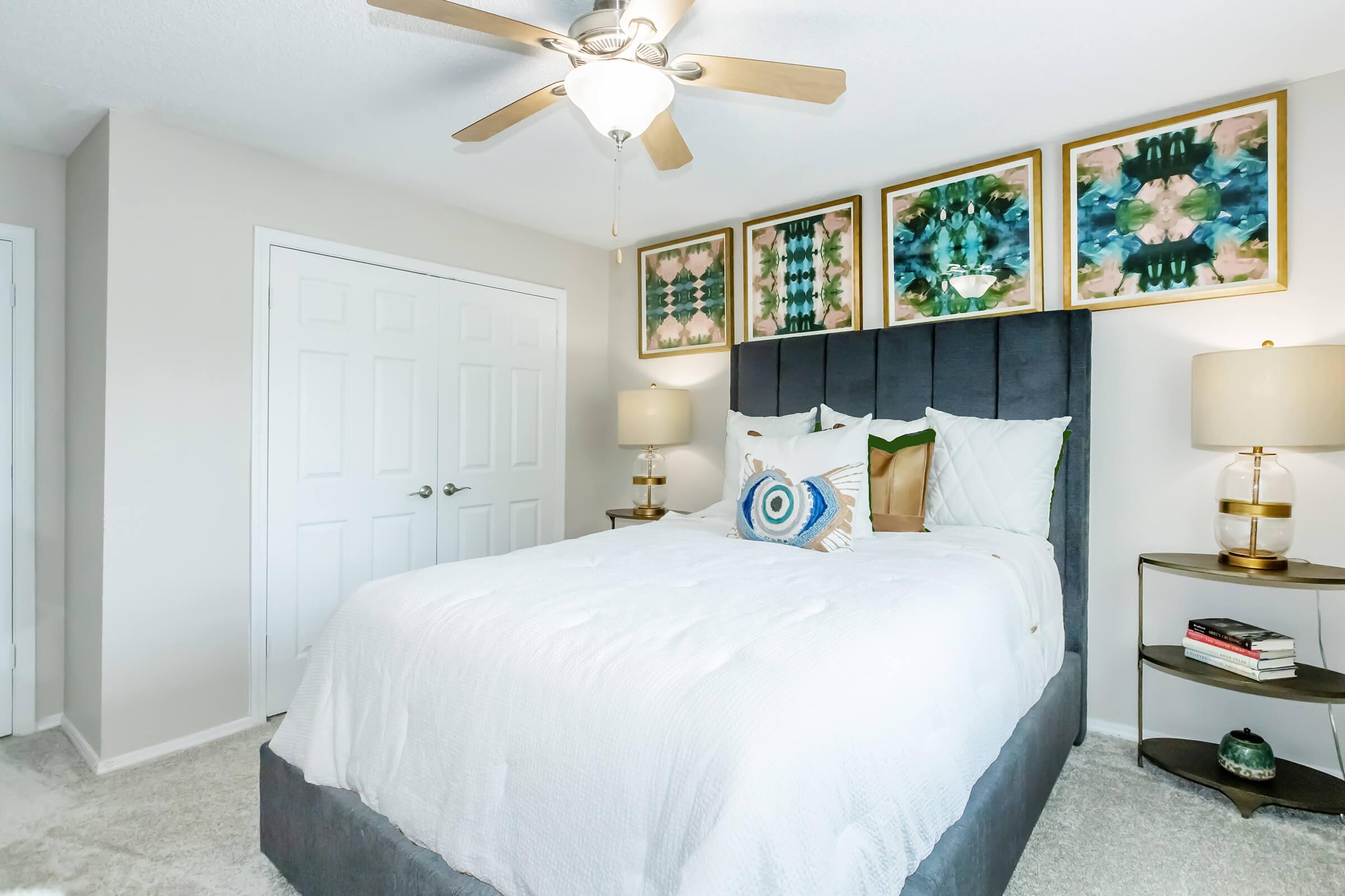 A cozy bedroom featuring a plush grey bed with decorative pillows and a quilt. Above the bed, there are four framed art pieces. On each side, stylish nightstands hold lamps and books. The room has a ceiling fan and light-colored walls, creating a welcoming atmosphere.