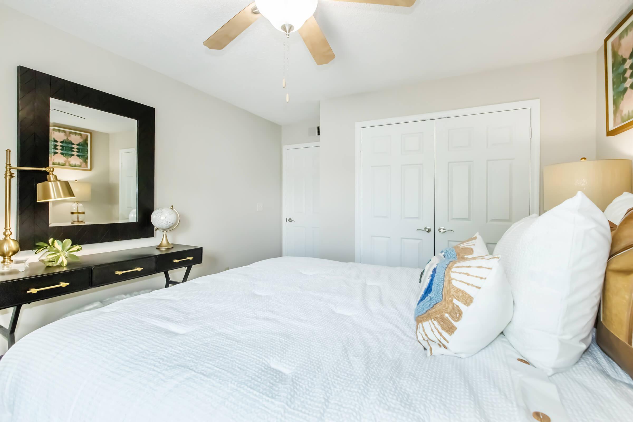 A cozy bedroom featuring a white bedspread with decorative pillows, a dark wood dresser with a lamp and decorative globe, and a large mirror. Soft lighting enhances the neutral-toned walls, creating a calm and inviting atmosphere. Two closet doors are visible in the background.