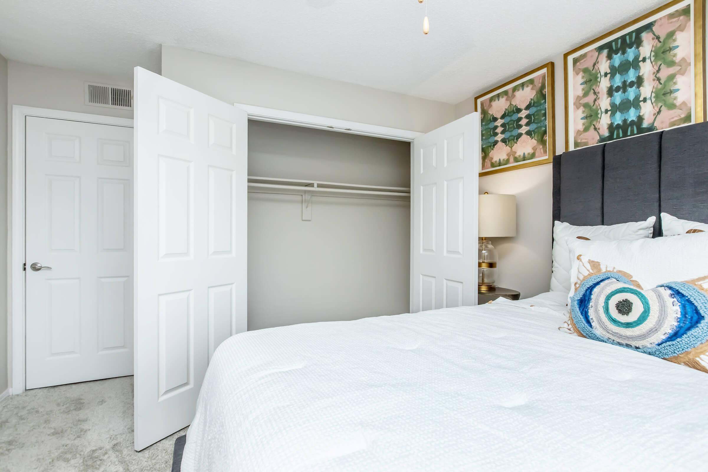 A neatly arranged bedroom featuring a bed with decorative pillows, a closet with sliding doors partially open, and framed artwork on the wall above the bed. The room has soft lighting and a carpeted floor, creating a cozy and inviting atmosphere.