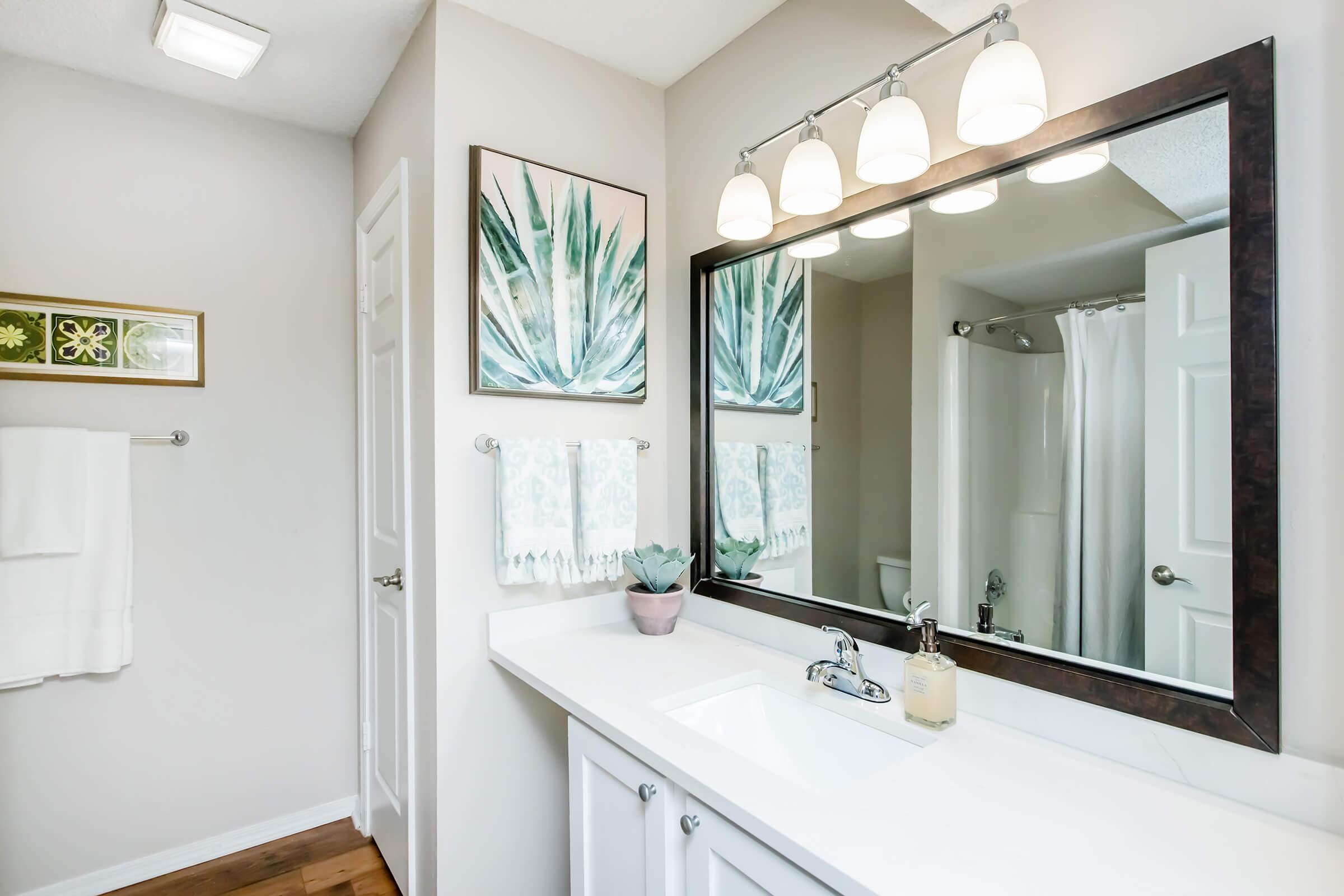 A modern bathroom featuring a white vanity with a sink, a large mirror with three light fixtures above, and decorative wall art depicting green plants. The space includes a shower area with a curtain, a towel rack with white towels, and a small potted plant on the countertop. The walls are painted in neutral tones.