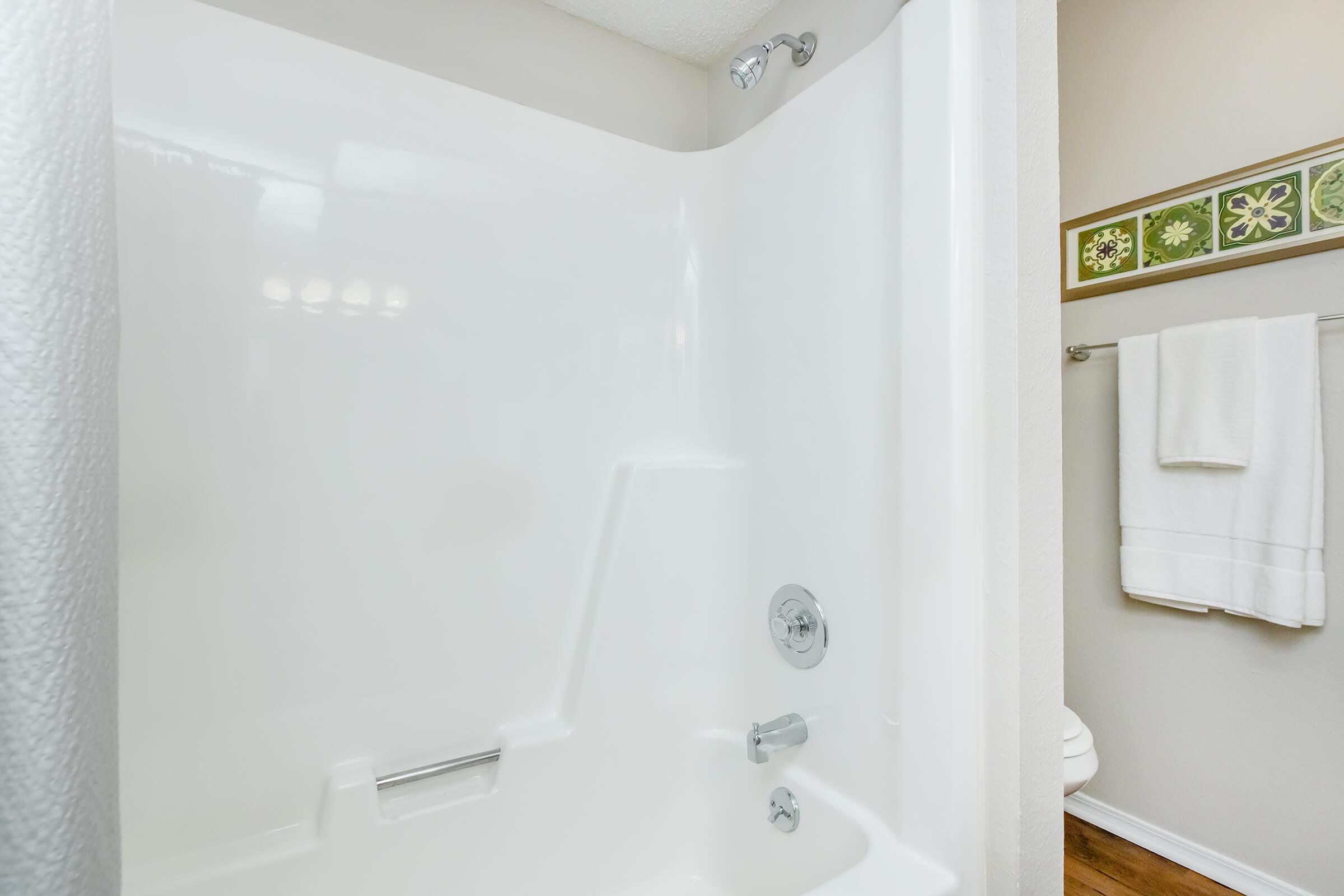 A clean and refreshingly simple shower stall with a white acrylic interior, a chrome showerhead, and a single lever control. The corner design maximizes space, while a towel rack with white towels is visible in the background, enhancing the bathroom's functionality and style.