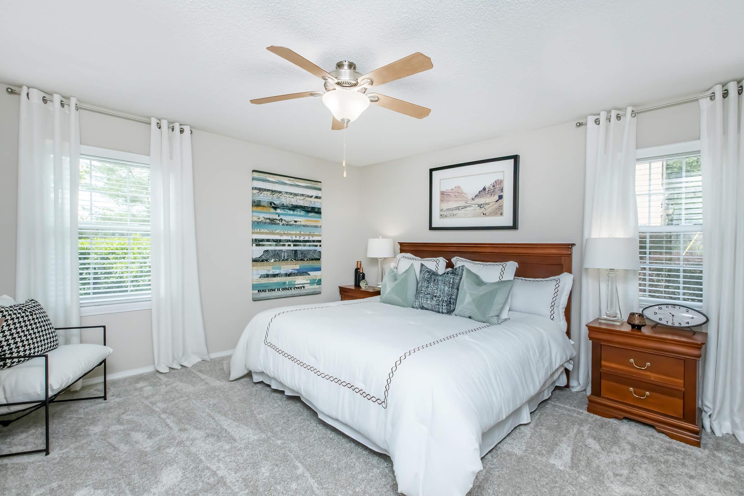 A cozy bedroom featuring a queen-sized bed with white bedding, decorative pillows, and a wooden headboard. The room is well-lit with natural light from two windows covered by sheer curtains. There’s a piece of wall art above the bed, a nightstand with a clock, and a chair in the corner, all set on soft carpet.