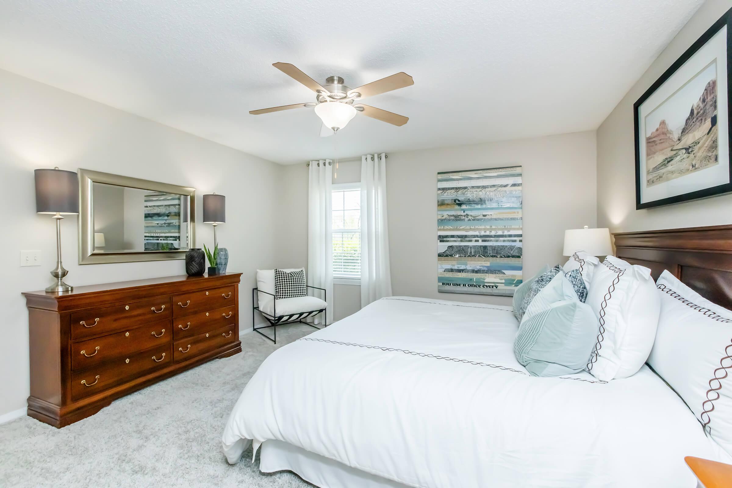 A well-decorated bedroom featuring a large bed with white bedding, a wooden dresser, two lamps, and a ceiling fan. There's a chair in the corner, and the walls are adorned with artwork. The room has light-colored carpet and bright natural light coming through the window.