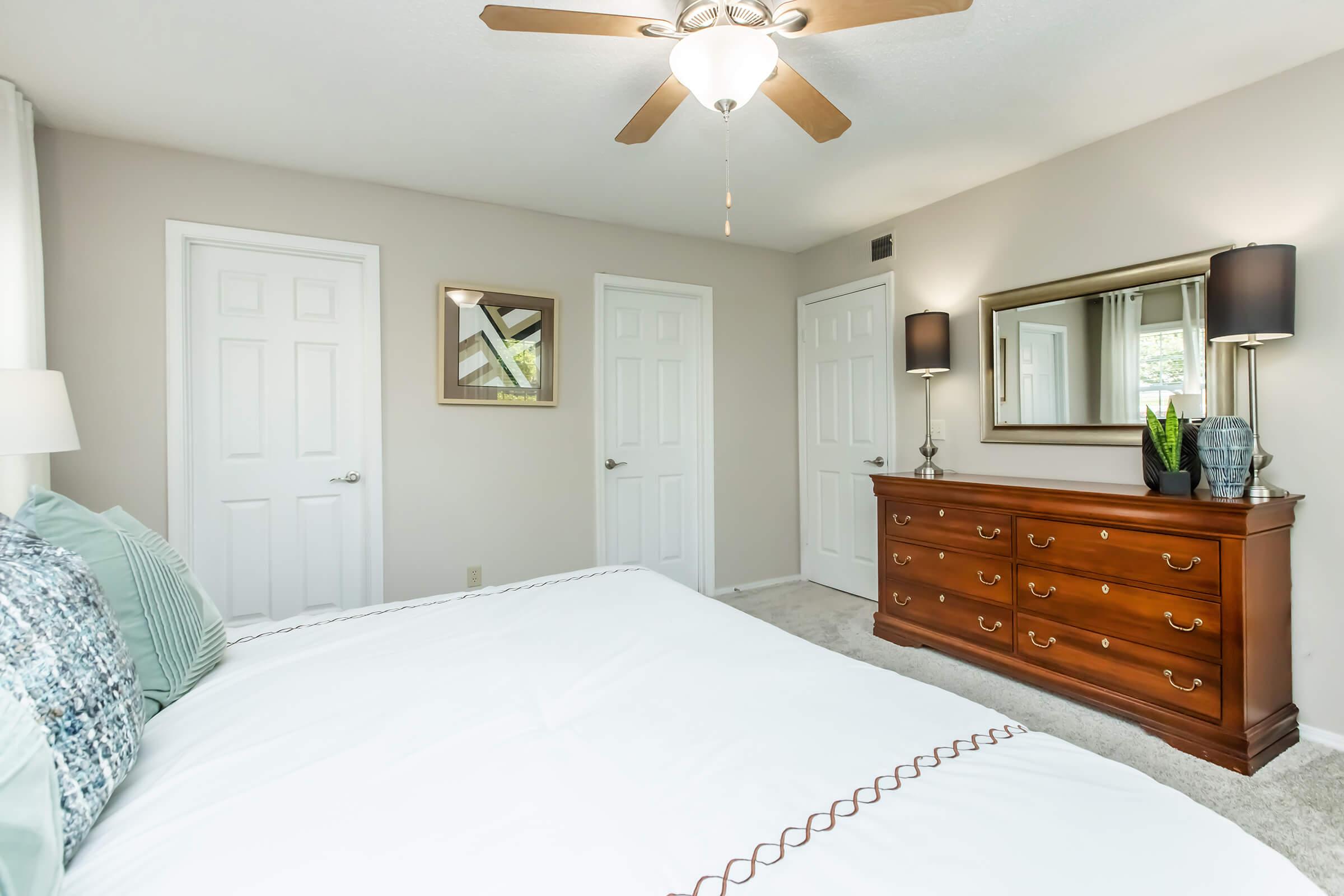 A well-decorated bedroom featuring a large bed with white bedding, a wooden dresser with multiple drawers, and a mirror. There are two doors, one of which is partly open, and a ceiling fan overhead. Soft natural light filters through the curtains, giving the room a cozy atmosphere.