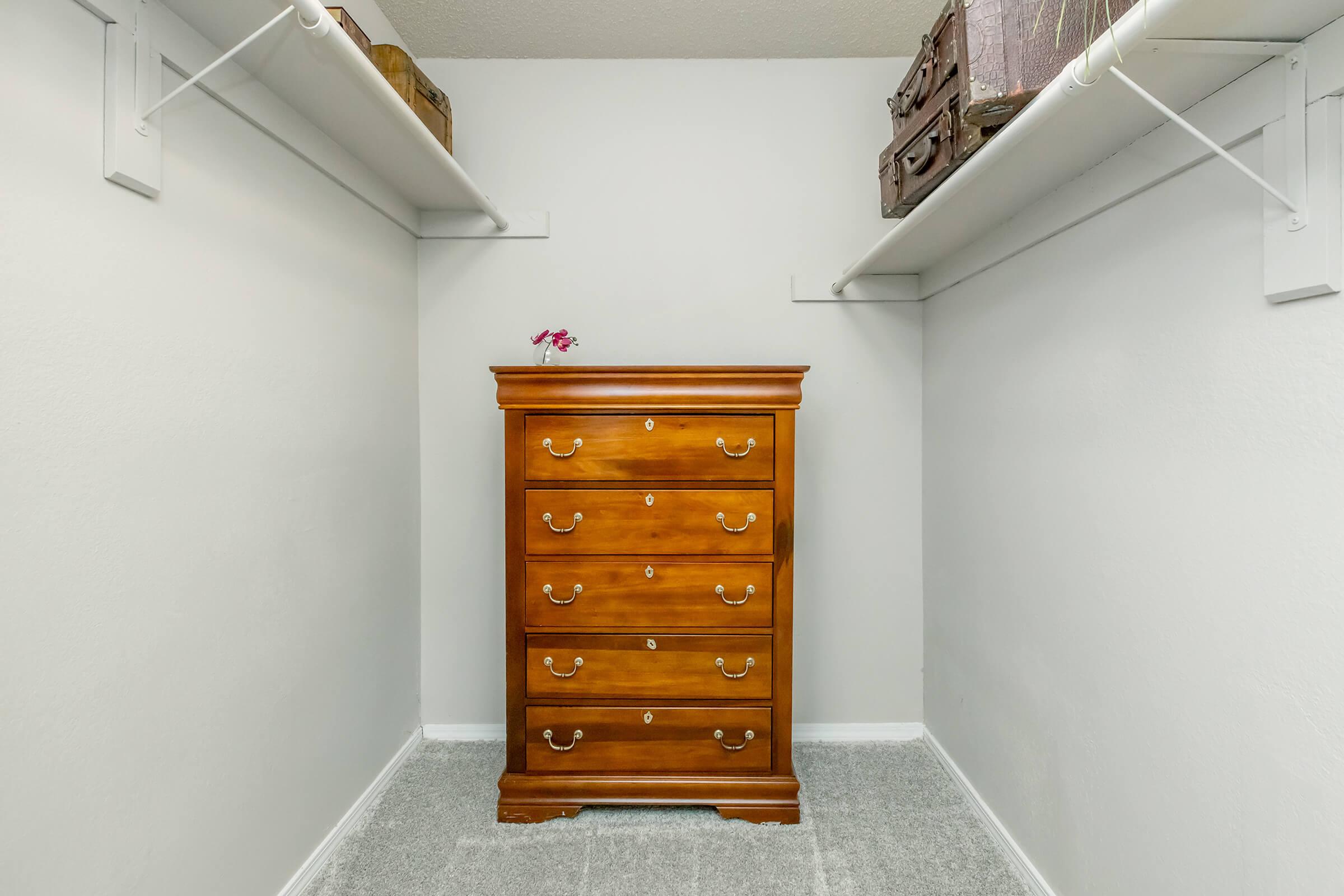 A neatly organized closet featuring a wooden four-drawer chest of drawers in the center. Above, there are two shelves with vintage suitcases on one side. The walls are painted white, and the floor is covered in light gray carpet. A small flower decorates the top of the dresser.