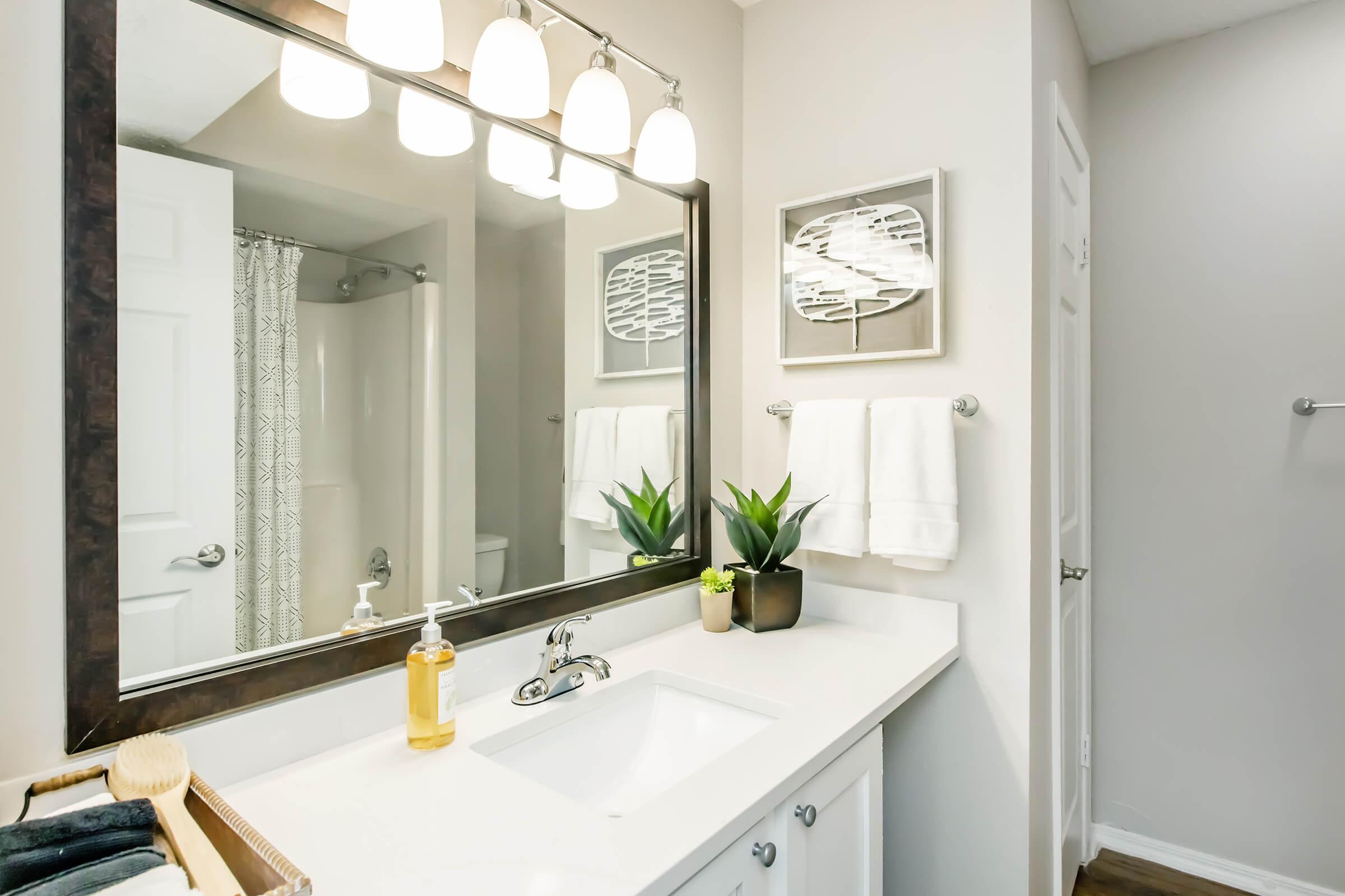 A clean, modern bathroom featuring a white countertop with a faucet, a large mirror with five light fixtures above, two white towels hanging on a towel rack, a small plant, and a bottle of soap. A patterned shower curtain is visible in the background, and the walls are painted in a light color.