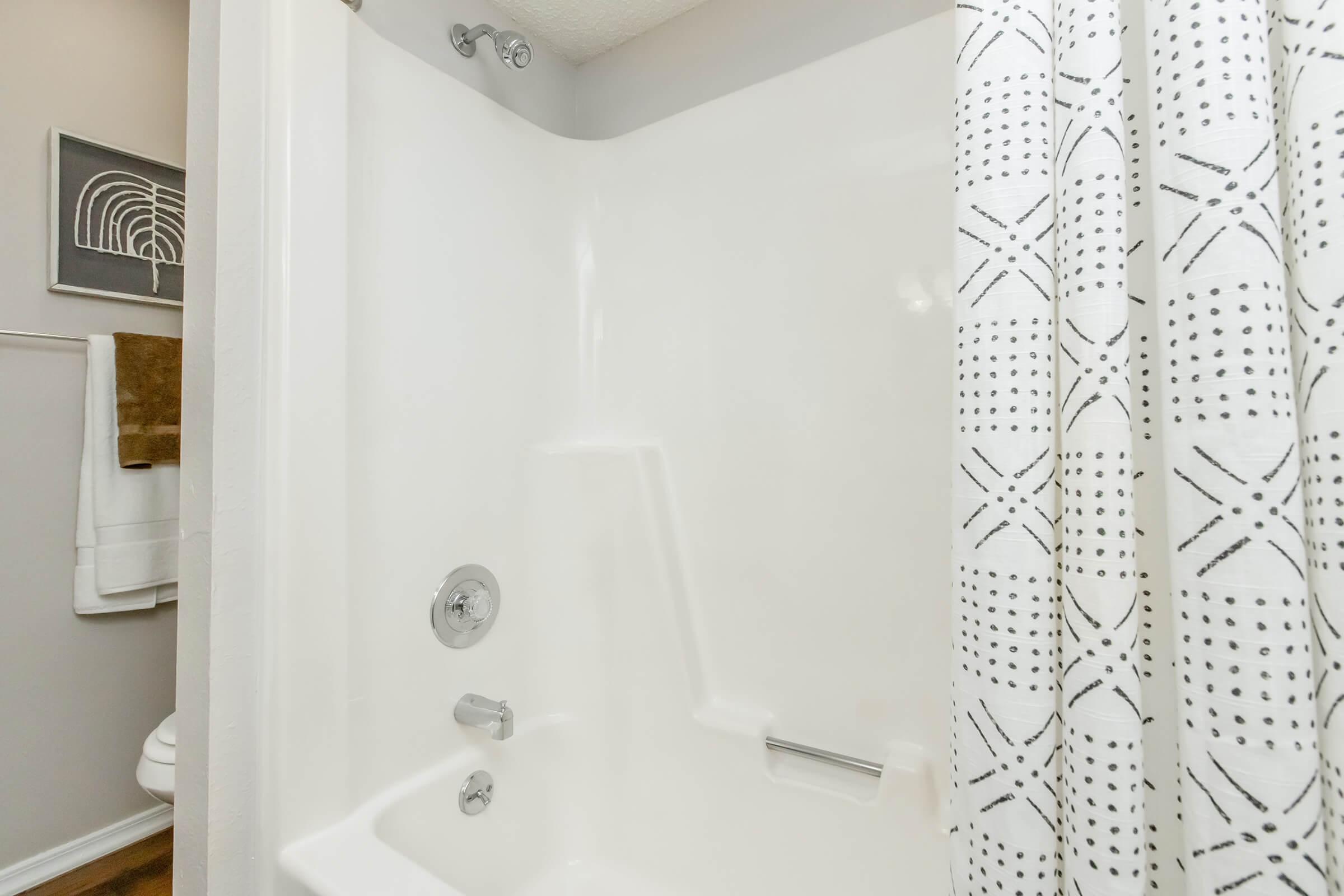 A clean, white shower space featuring a smooth, curved wall and a modern chrome showerhead. The image includes a shower curtain with a geometric black and white pattern, neatly arranged towels hanging nearby, and a glimpse of bathroom fixtures in the background.