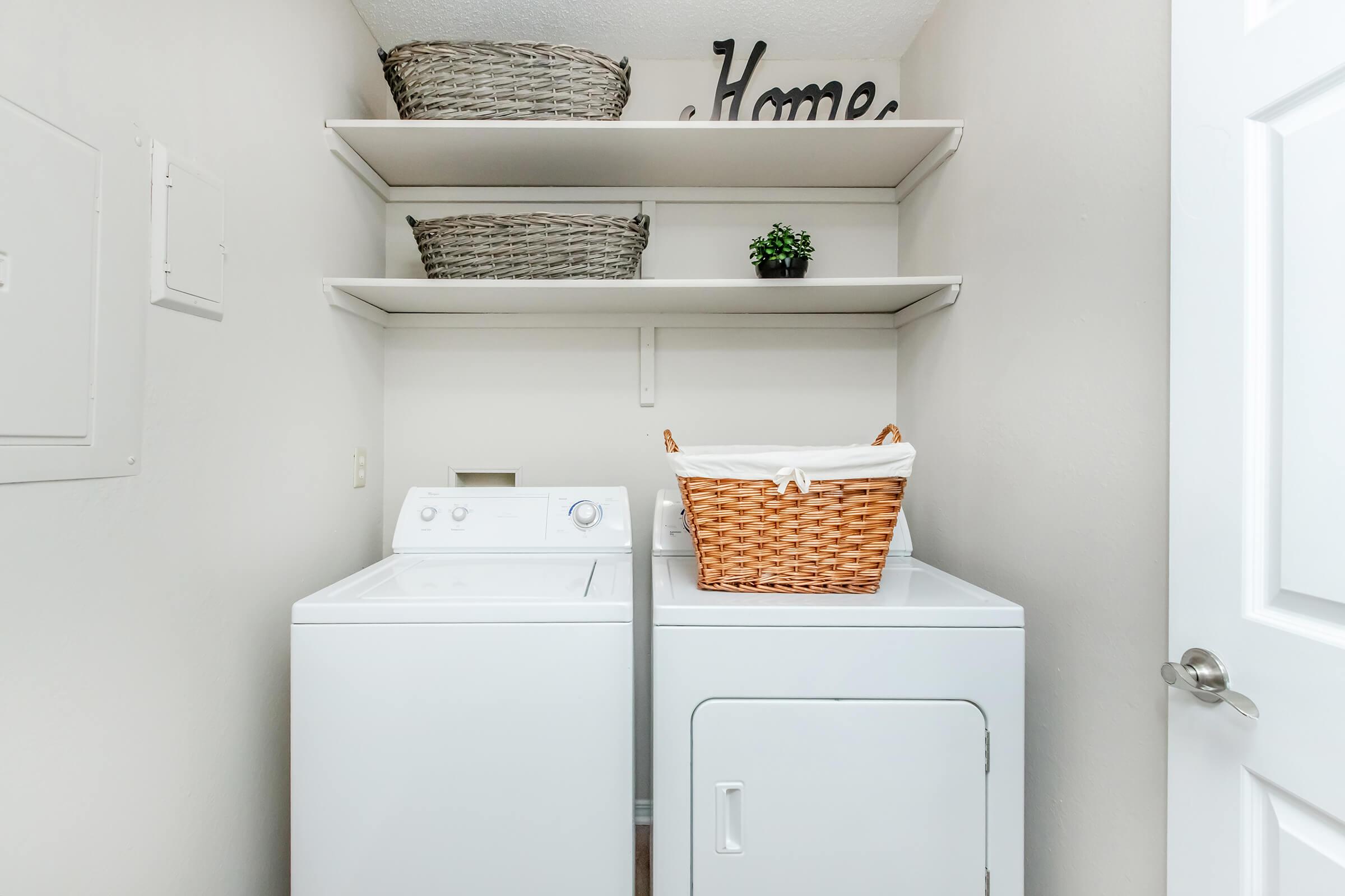 A laundry room featuring a white washing machine and dryer side by side. Above, there are two shelves with woven baskets and a small plant. A decorative piece that reads "Home" is placed on the top shelf. The walls are light-colored, creating a bright and organized space.