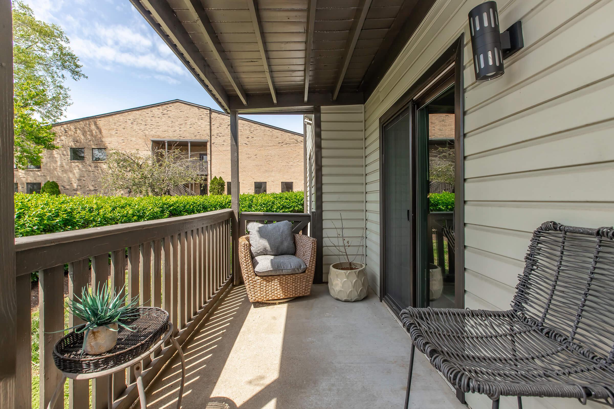 A cozy balcony with a wicker chair and a small table featuring a potted plant, overlooking a garden area. The space is well-lit by natural light, with a view of a neighboring building and green foliage in the background.