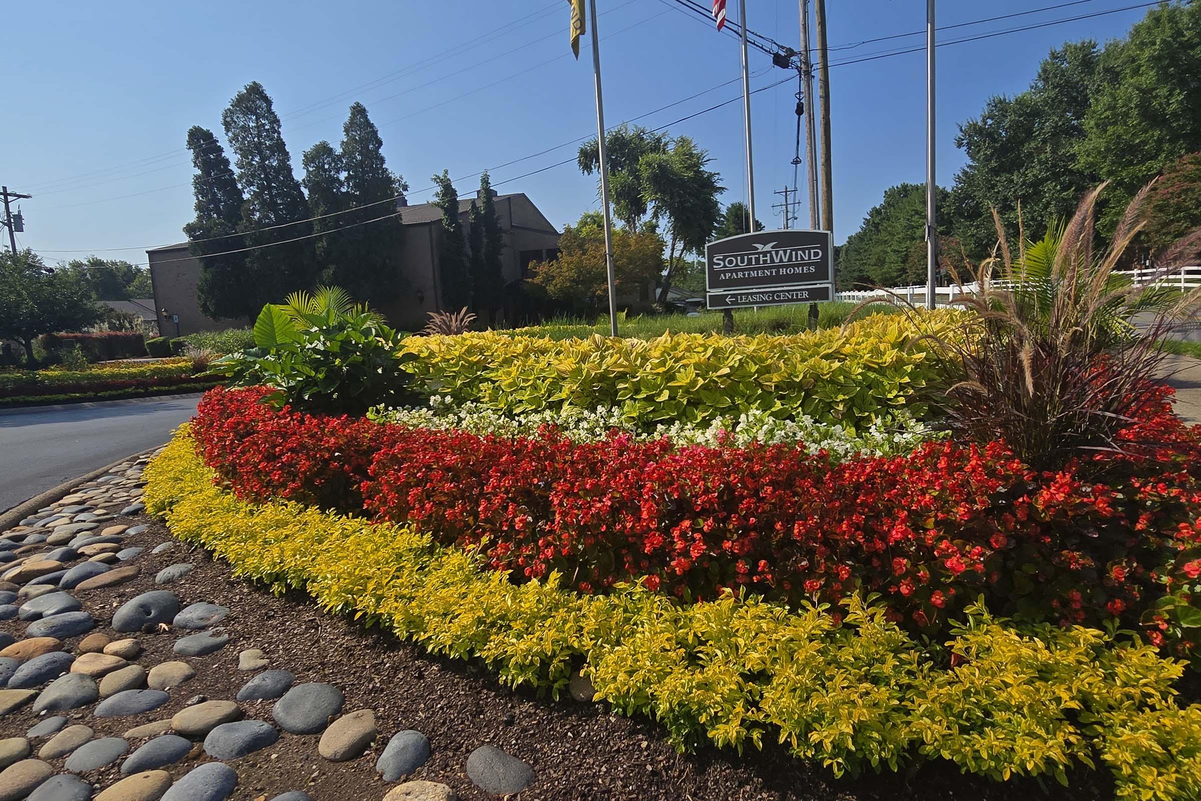A vibrant flower bed featuring red, yellow, and white flowers, bordered by smooth gray stones. In the background, a sign for Southwind Apartments is visible alongside trees and a clear blue sky. The scene is well-maintained, showcasing a welcoming entrance to the property.