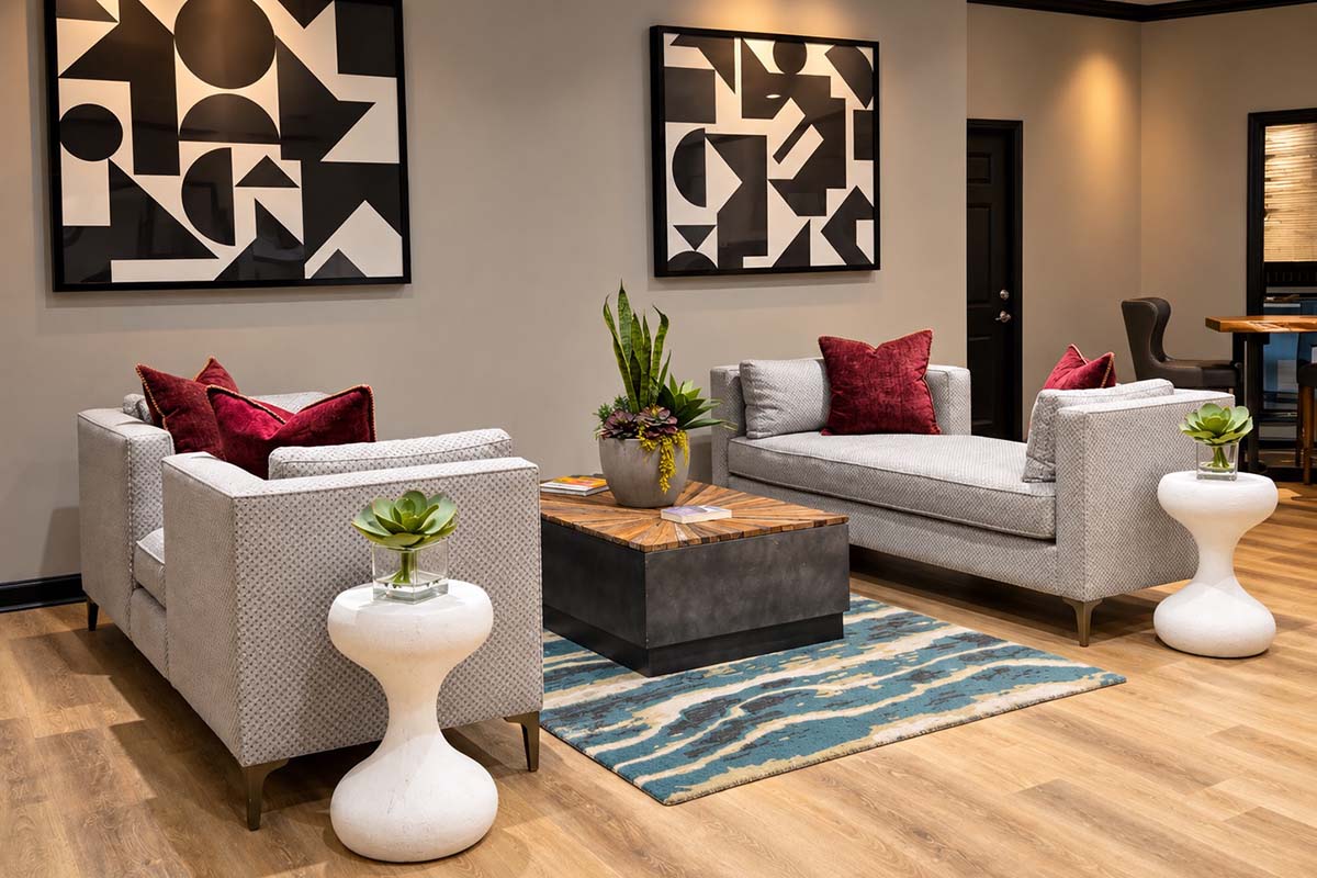 Contemporary lounge area featuring two stylish gray sofas with red accent pillows, a coffee table with a wooden top and dark base, and decorative plants in white pedestals. The walls are adorned with modern abstract artwork, and the flooring is light wood. A textured area rug adds warmth to the space.