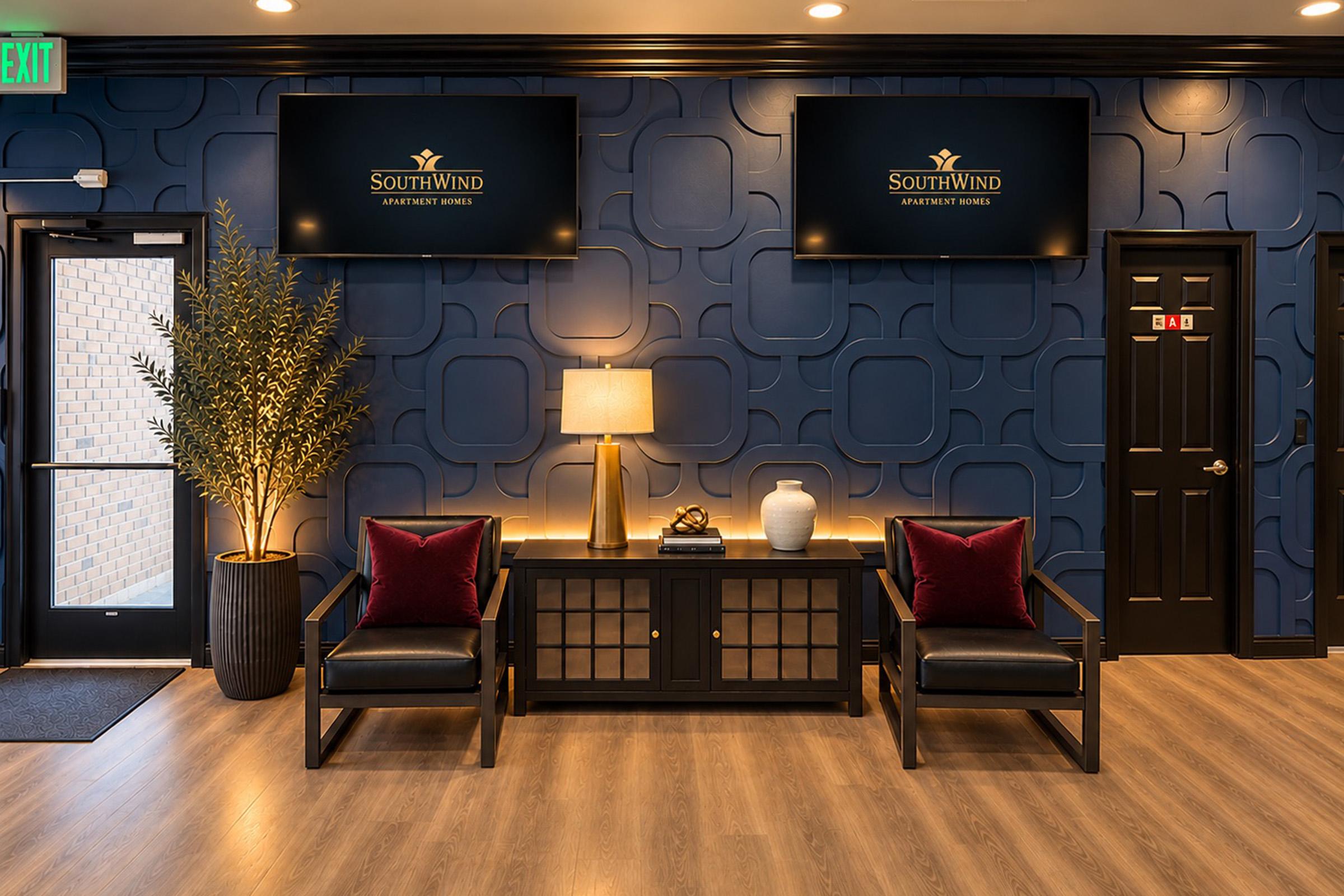 A modern lobby area featuring a dark blue wall with textured panels. Two black armchairs with red pillows are positioned beside a wooden cabinet, a table lamp, and a decorative vase. Two screens display the "SouthWind Apartment Homes" logo. An exit door is visible on the left.