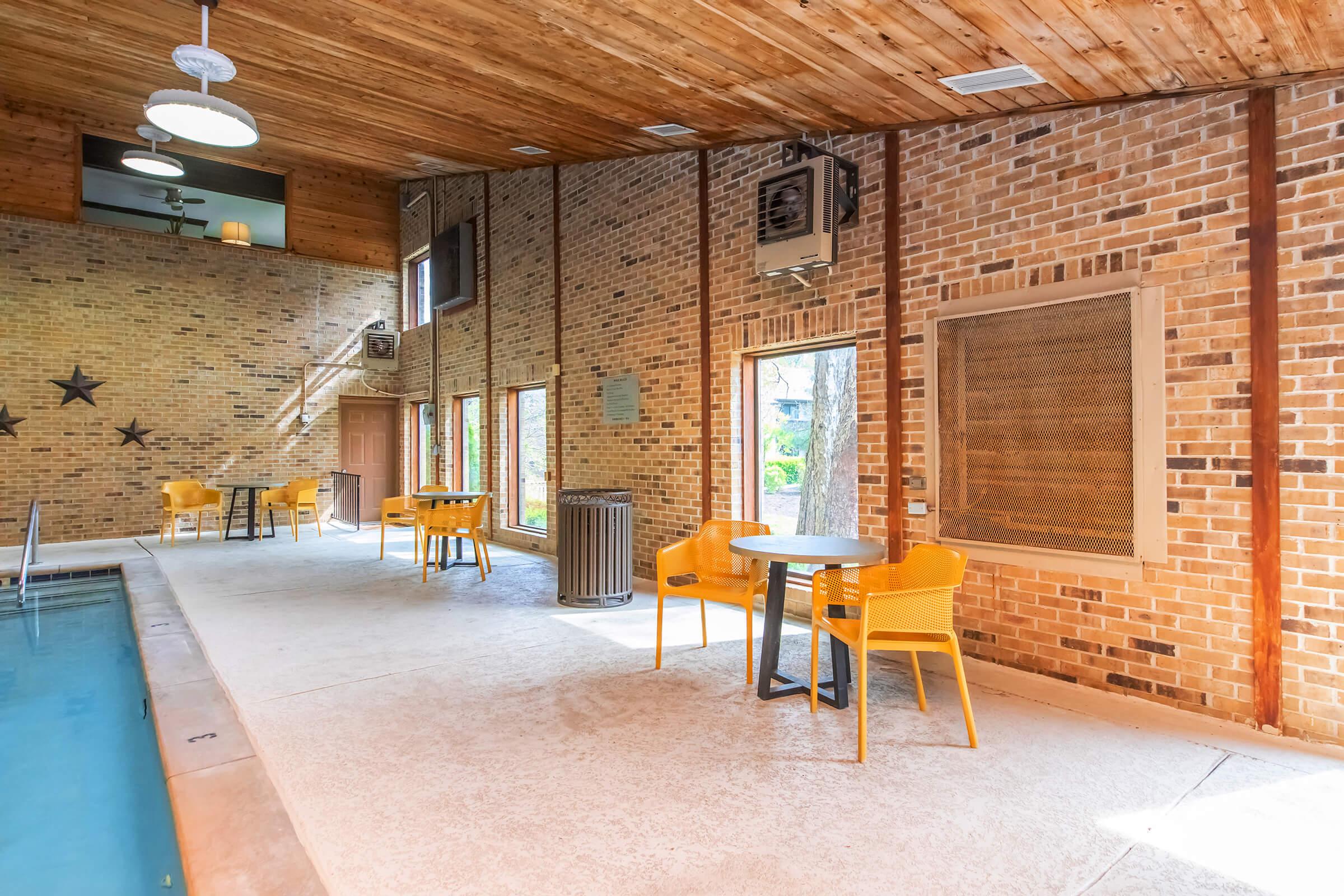 Indoor pool area with a natural wood ceiling, brick walls, and large windows letting in natural light. There are a few yellow chairs and tables, a swimming pool on one side, and decorative stars on the wall, creating a warm and inviting atmosphere.