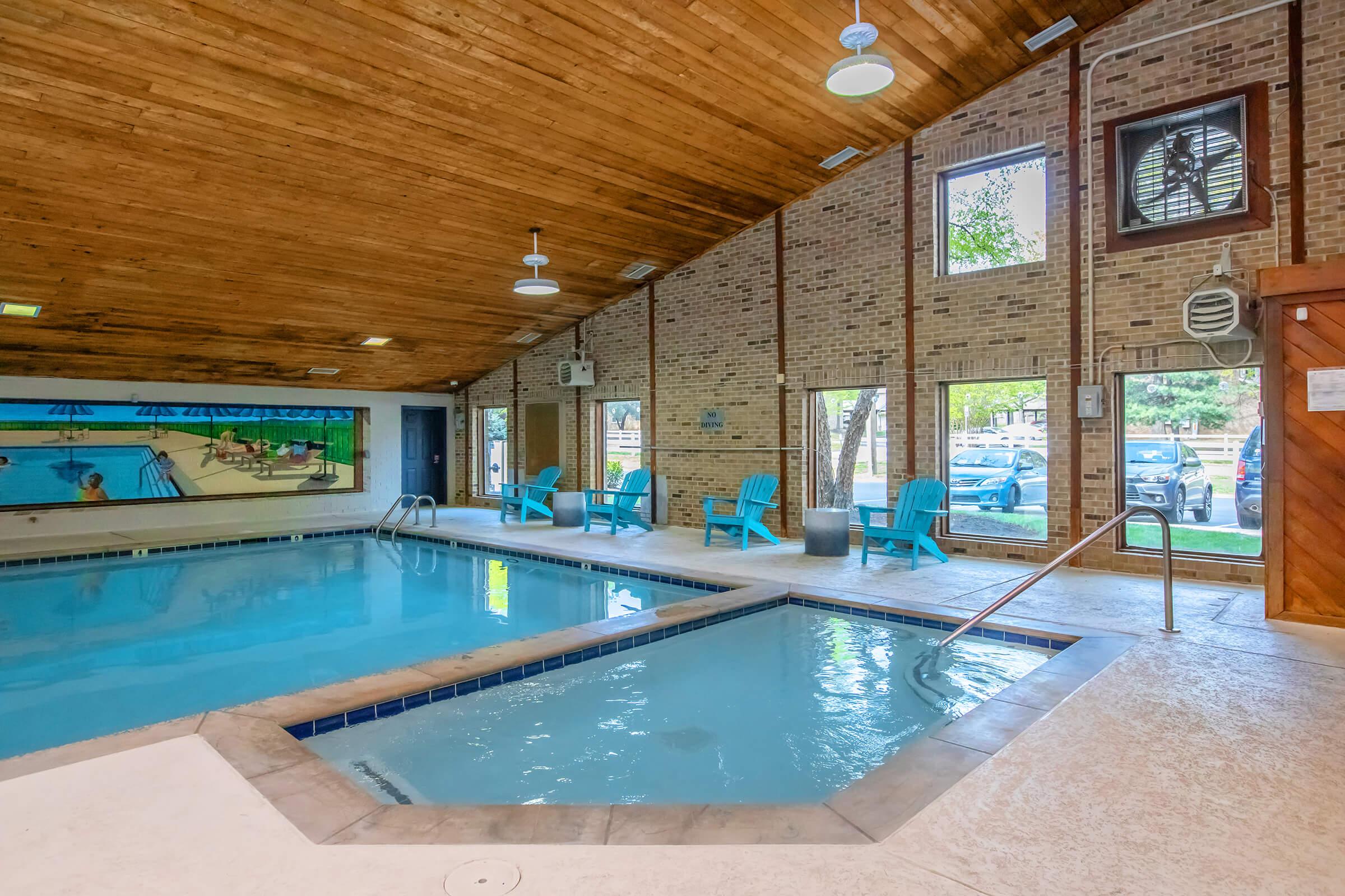Indoor swimming pool area featuring a main pool and a smaller hot tub, surrounded by wooden ceiling beams and brick walls. Large windows allow natural light, showcasing blue chairs and a mural depicting a beach scene. Nearby, vehicles are visible through the windows.