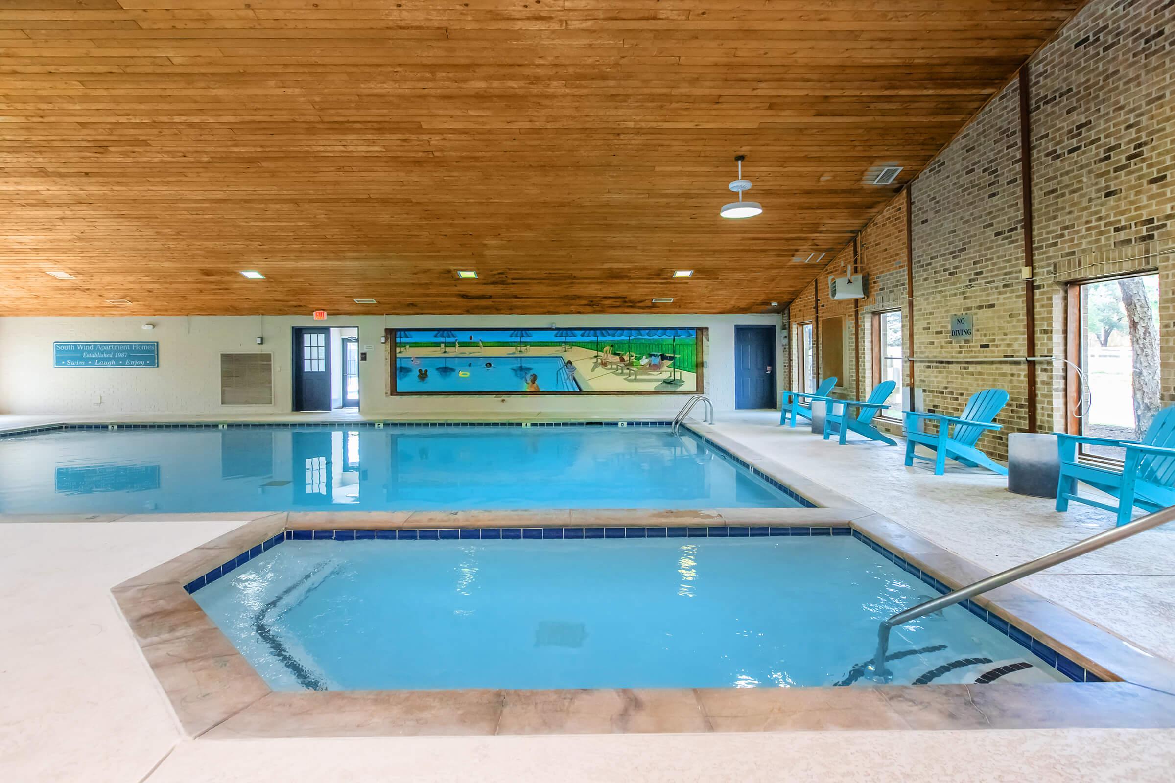 Indoor pool area featuring a large swimming pool and a smaller hot tub. The ceiling has wooden beams, and there's natural light coming through large windows. Colorful murals depicting aquatic scenes are on the walls, and there are blue adirondack chairs for lounging nearby.
